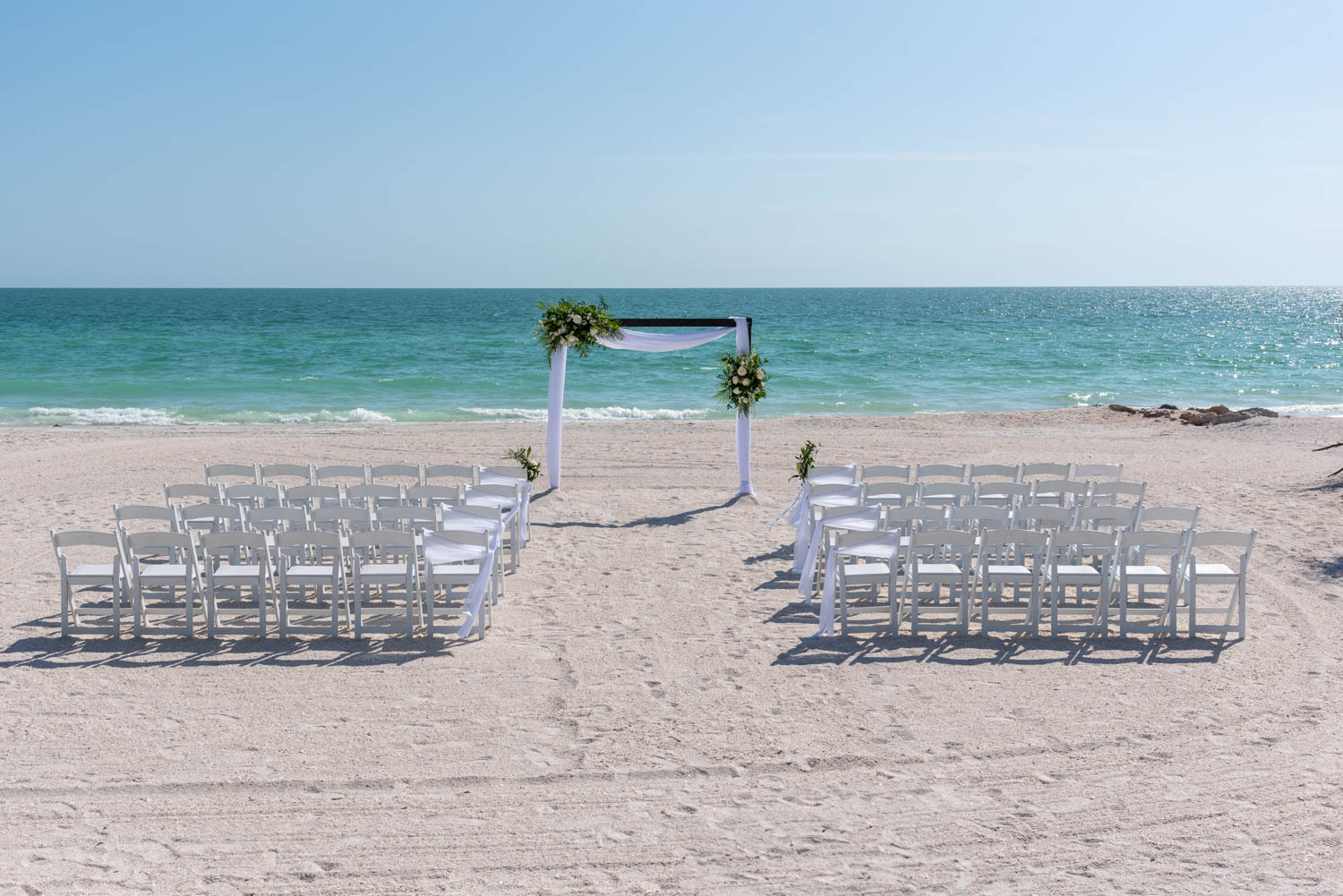 beach wedding venue set up with white chairs and arch with flowers