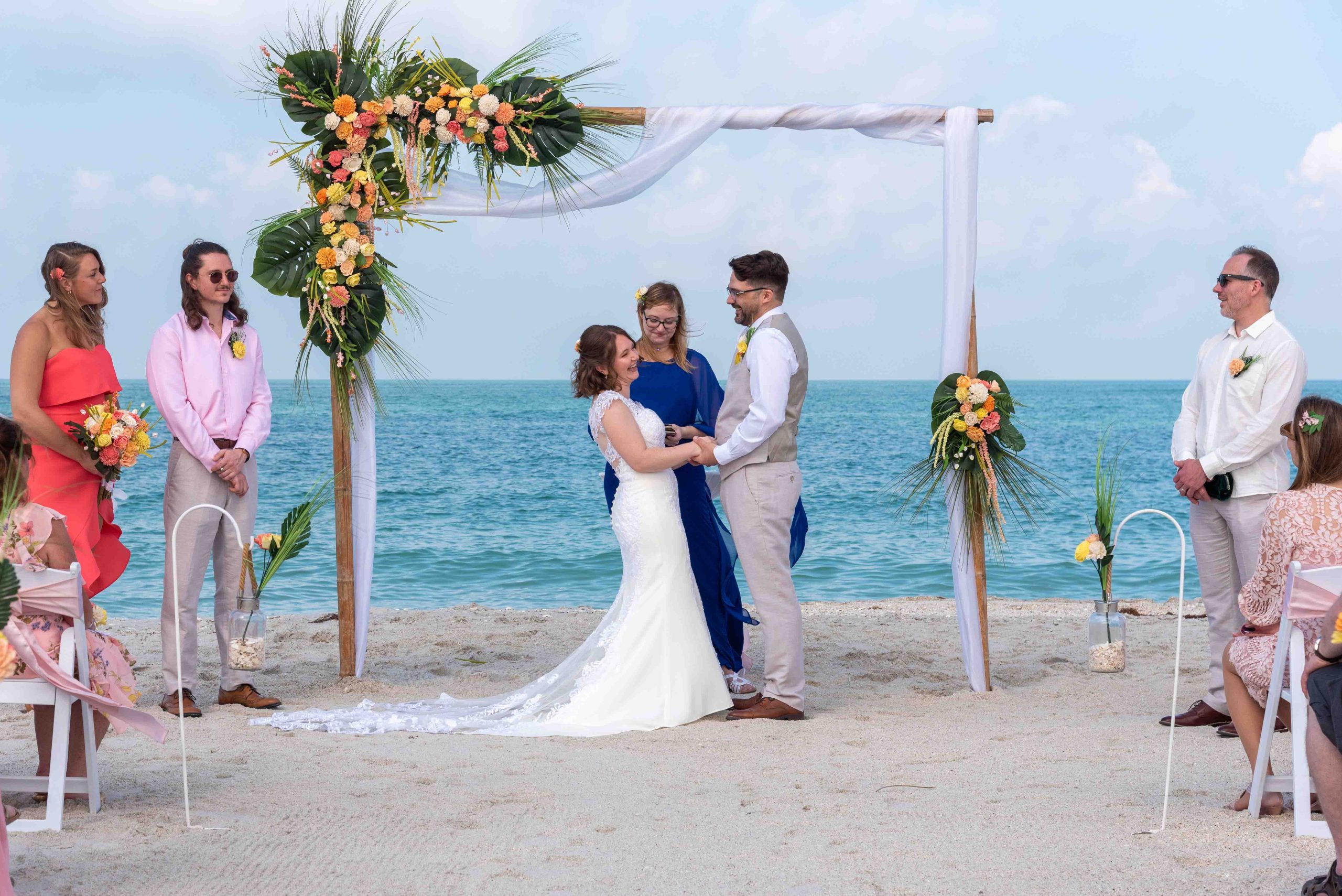 wedding ceremony on the beach with the bride, groom and officiant and beautiful blue water behind them