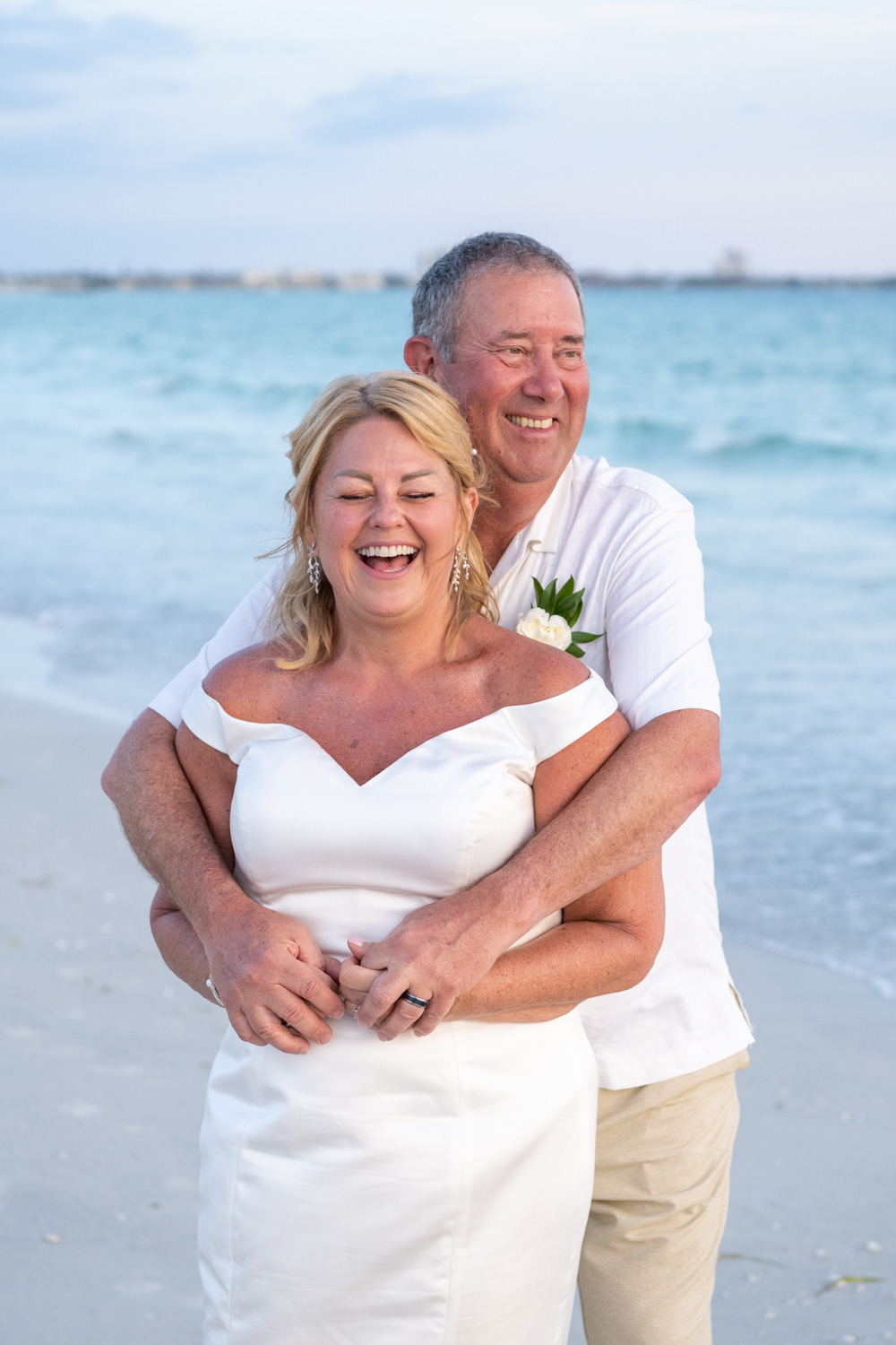 bride laughs and smiles as husband holds her from behind while on the beach with blue water behind them