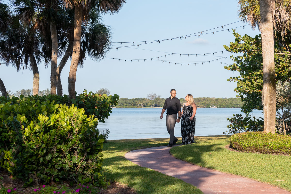 couple hold hands as they walk along the brick sidewalk with the water in the background