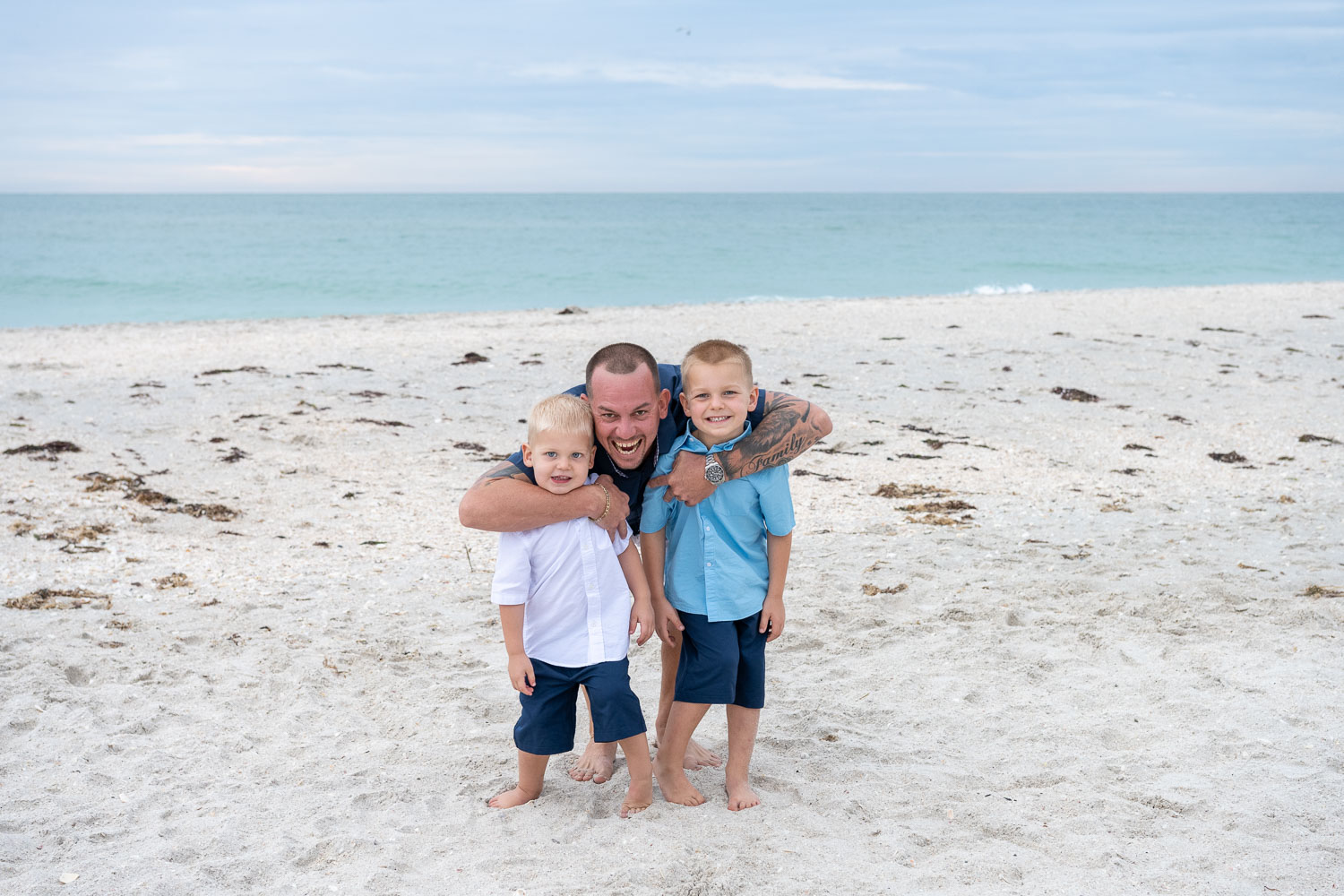 father leans down and wraps his arms around his two young sons as they stand on the beach
