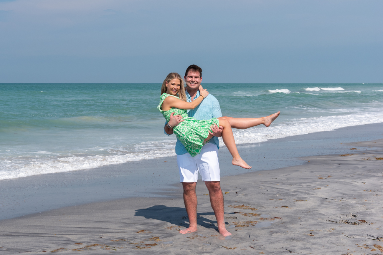 man lifts fiance in air as they stand on the beach on Palm Island