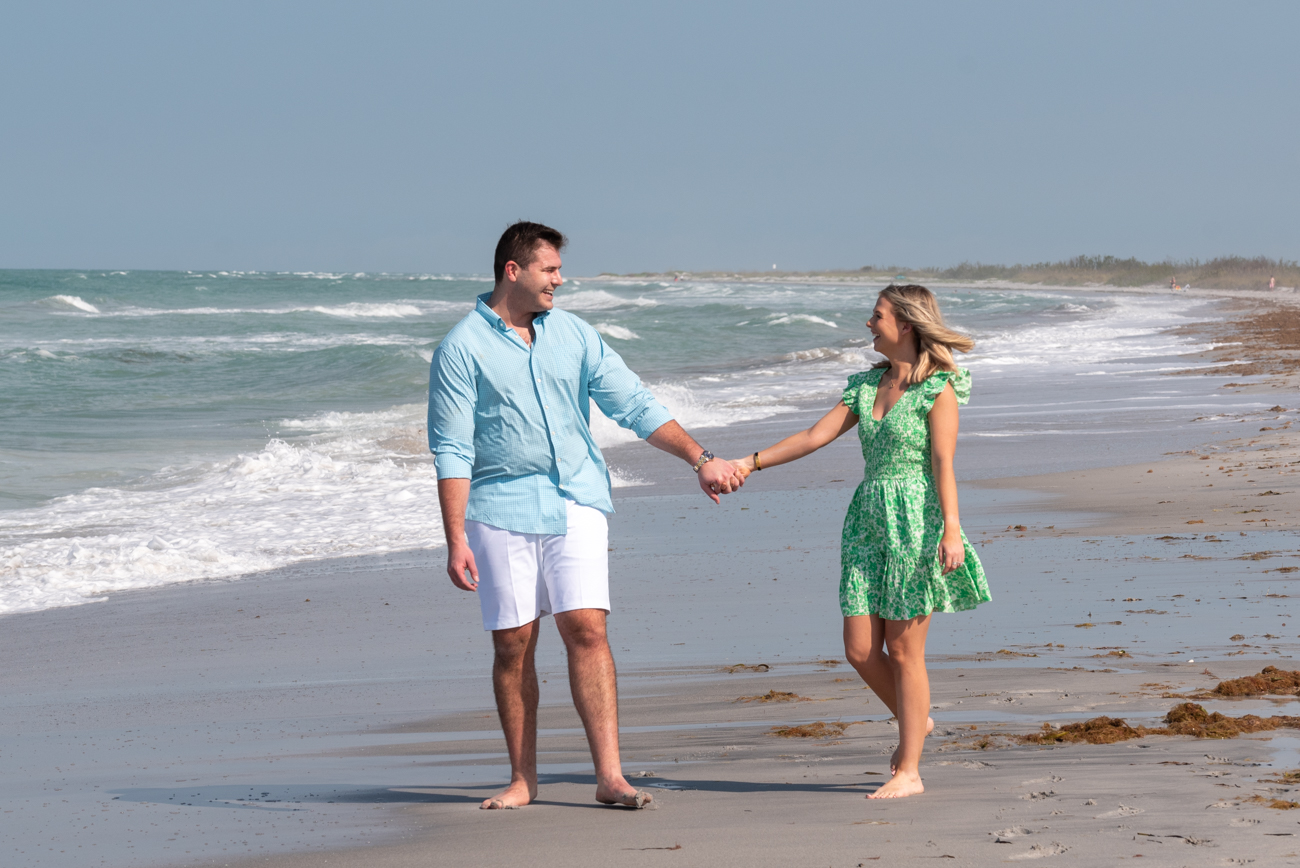 newly engaged couple hold hands as they look at each other on the beach