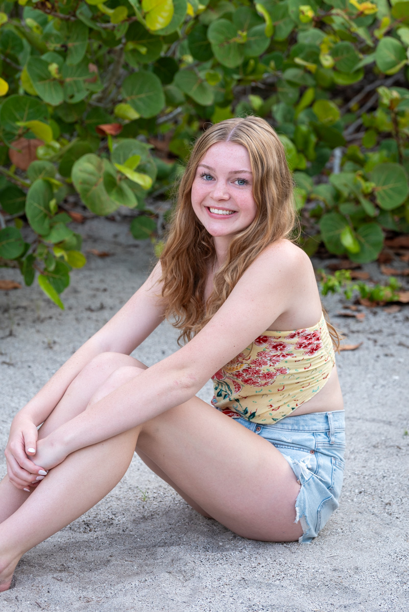 girl sitting on sand wrapping her arms around her legs with sea oats behind her