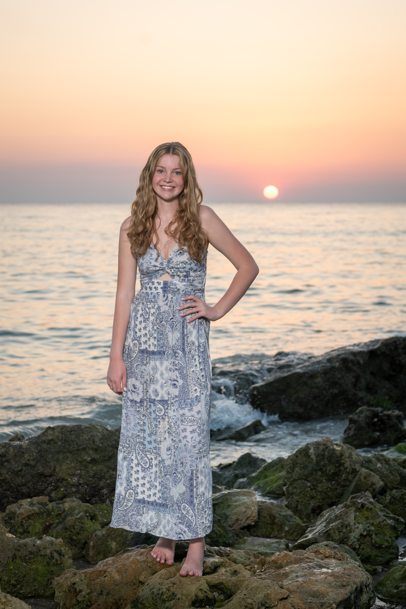 senior photo at Caspersen Beach in Venice with girl dressed in a purple dress standing on the boulders as the sun sets behind her
