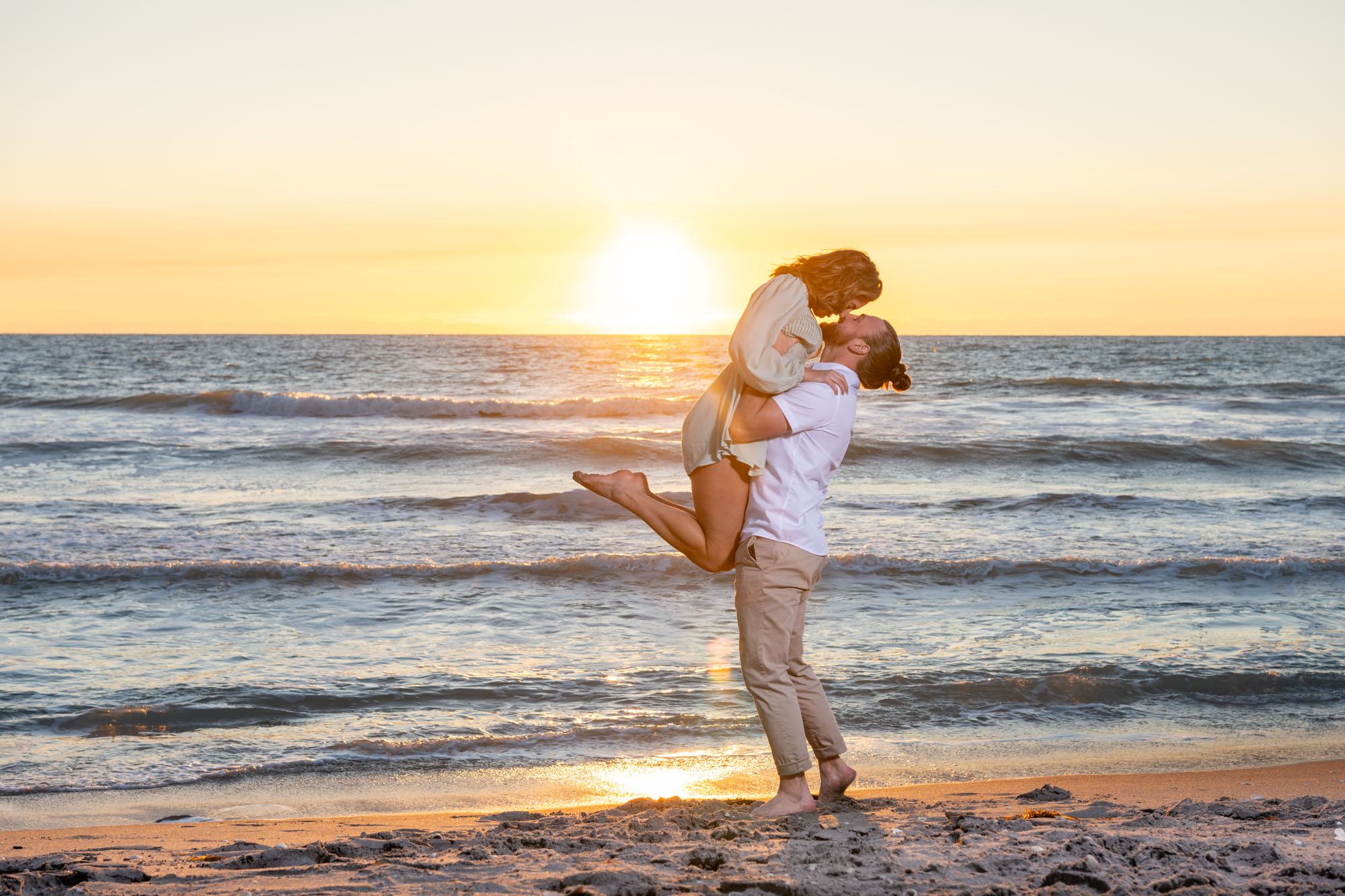 guy lifts girl up over his head as the sun starts to set behind them as they stand on the beach