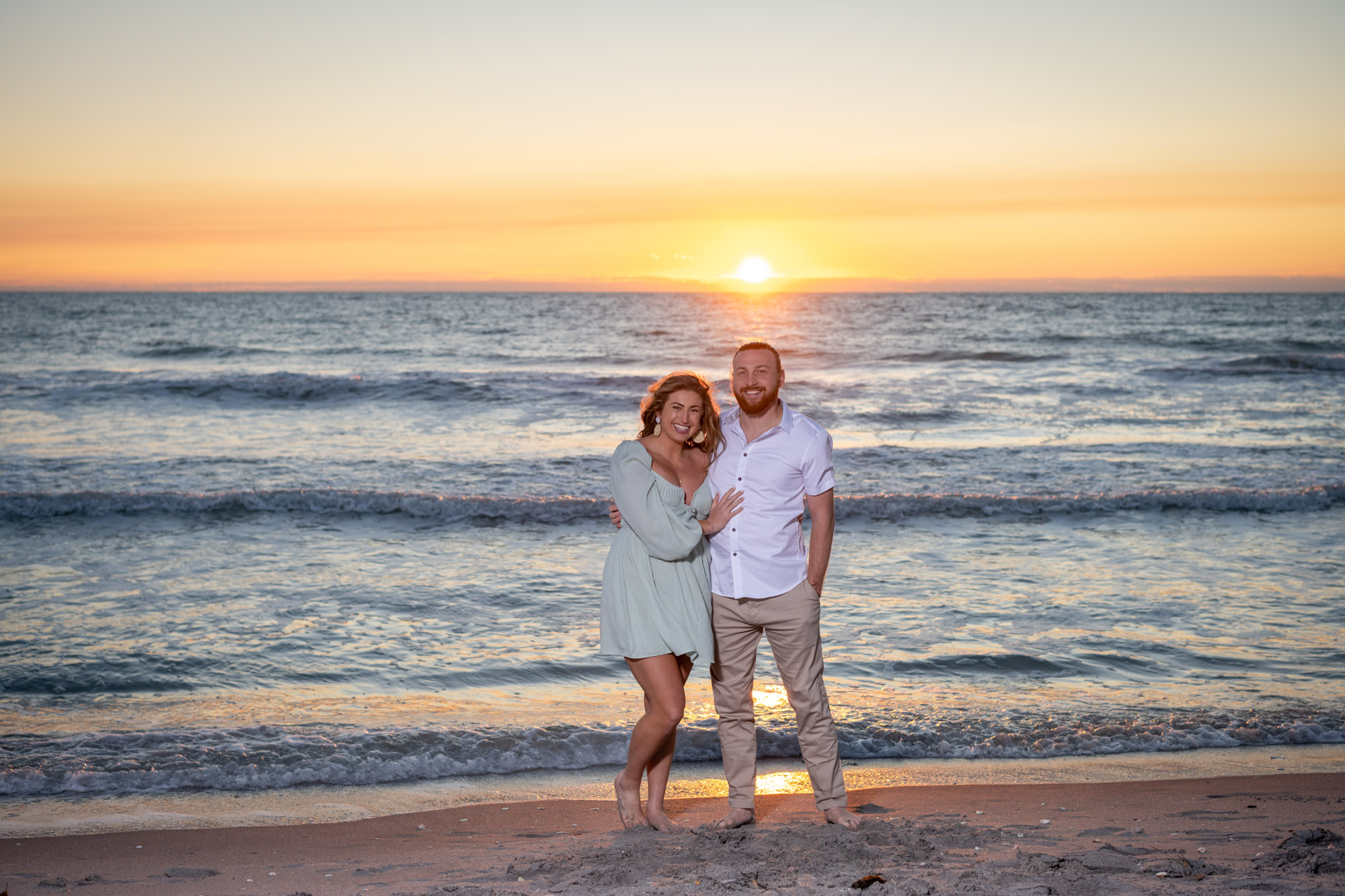 newly engaged couple stand on the beach as the sun sets behind them