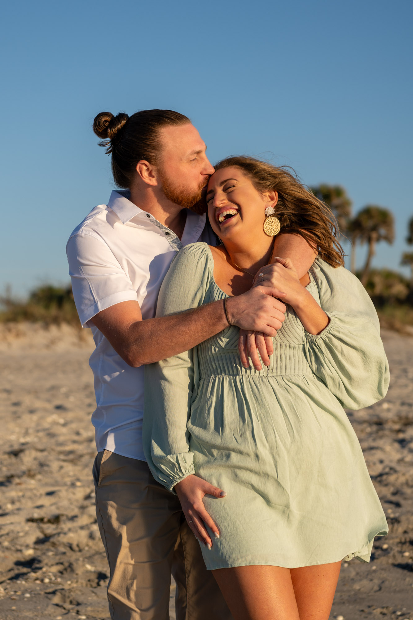 guy kisses girlfriend's forehead as she laughs with the sand and palm trees at Englewood beach behind them