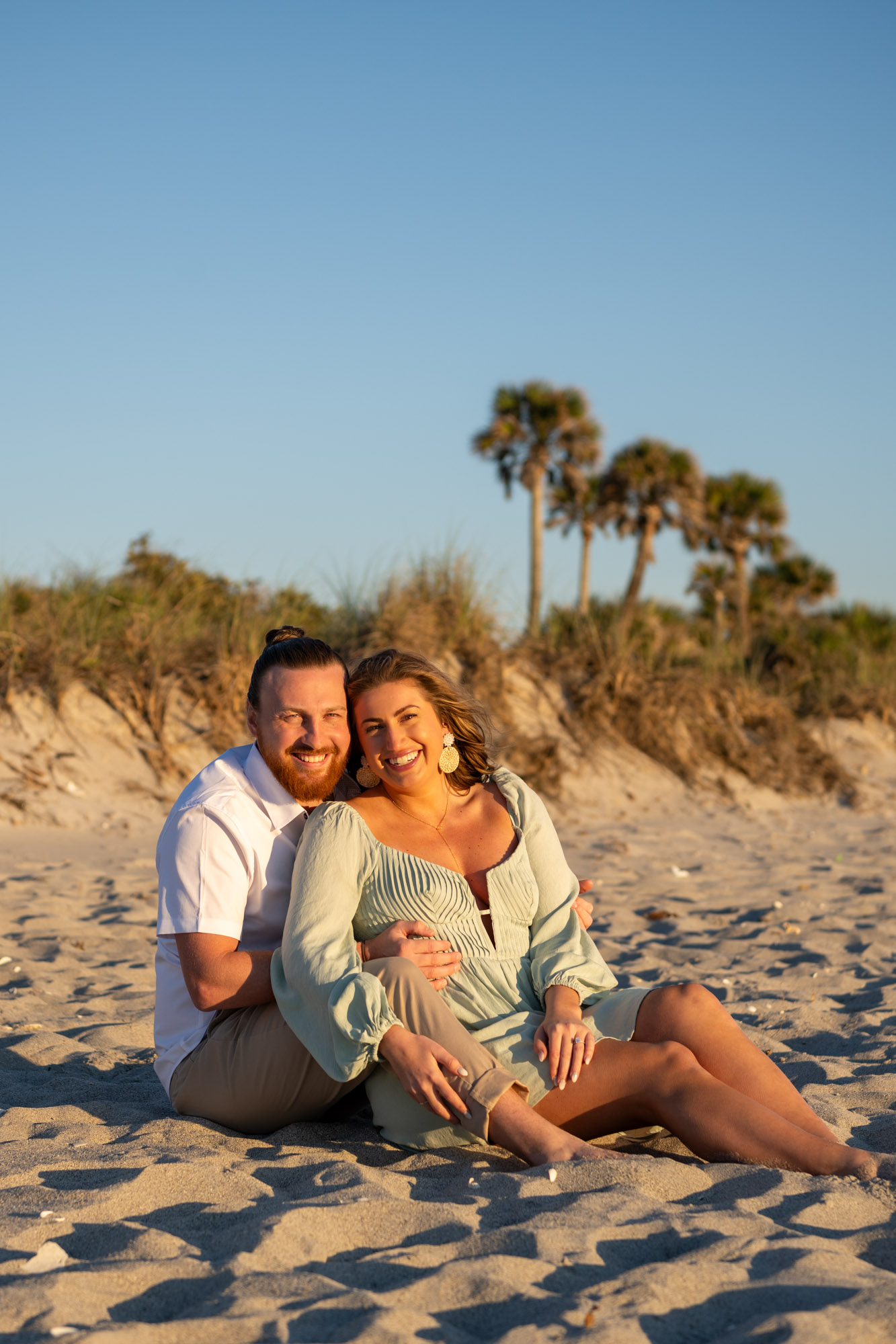 engaged couple sit in the sand at Englewood beach with palm trees behind them