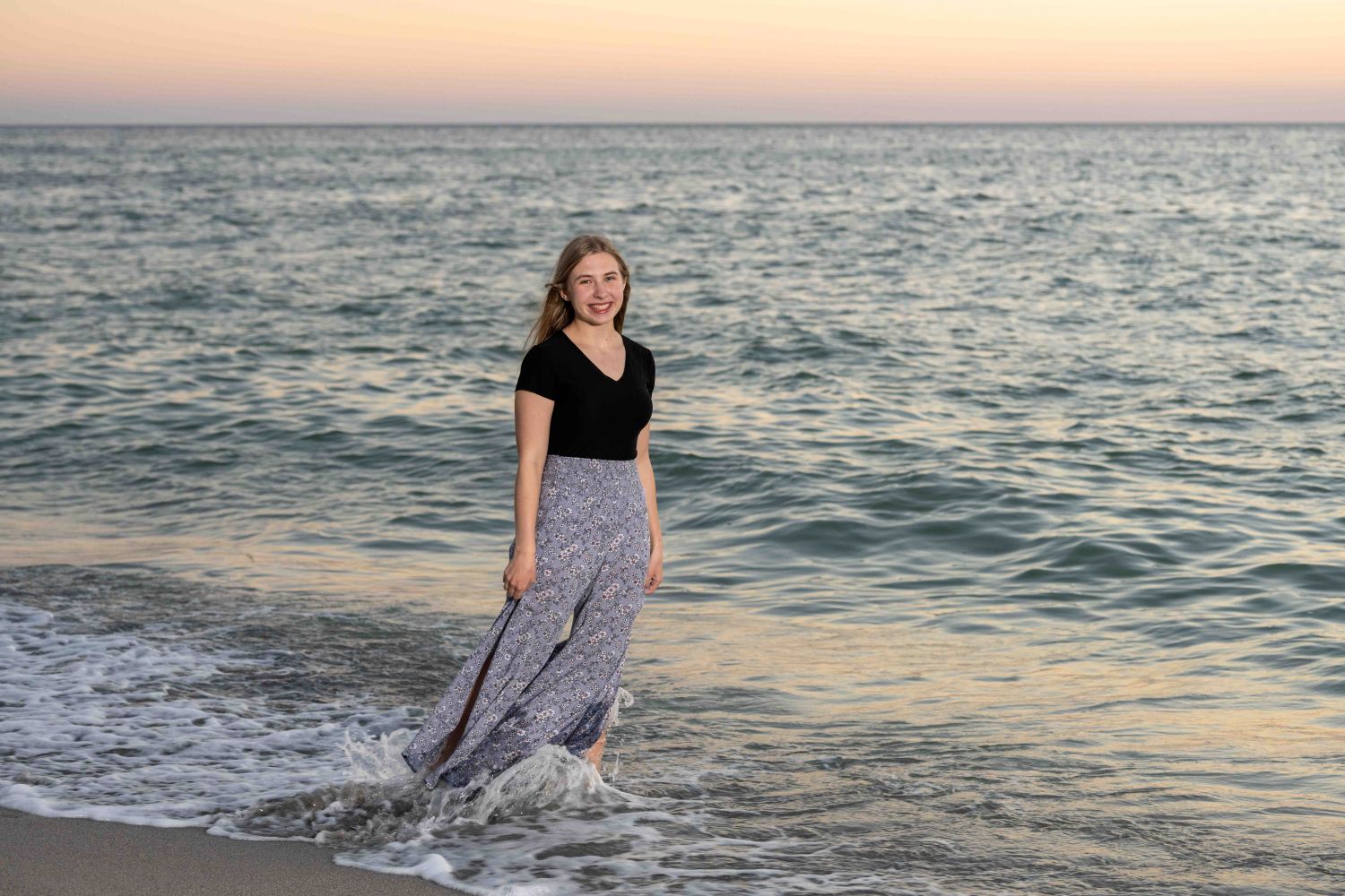 Senior photo of girl in black top and flowing skirt standing in water which rushes her feet as the golden hour soft sun is behind her