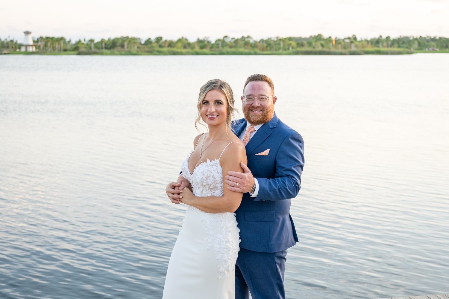 groom holds his bride from behind as they stand in front of the pond and lighthouse in Wellen Park