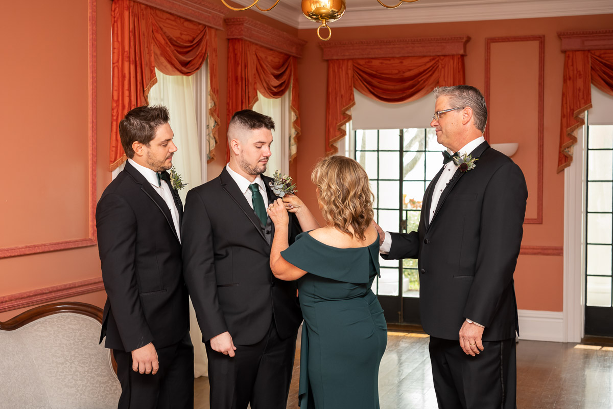 the groom's family stands inside as the guests arrive and his mother attaches his boutonniere