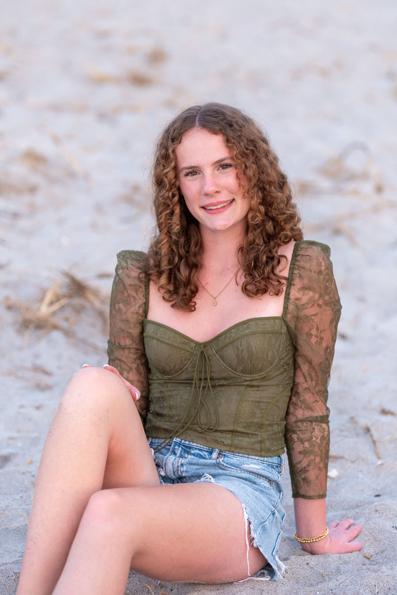 senior beach session with girl in green top and blue jean shorts sitting on the beach