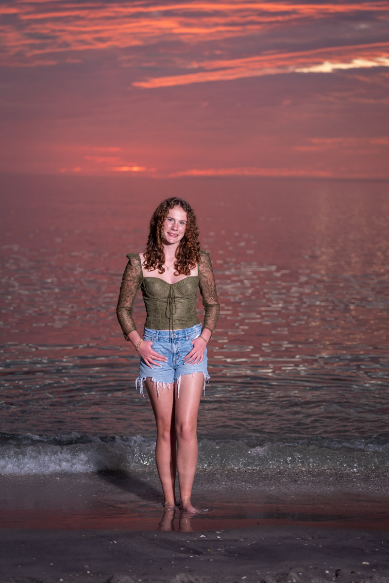 senior session on the beach with the sky turning bright pink after sunset and a girl in green top and blue jeans shorts by the water