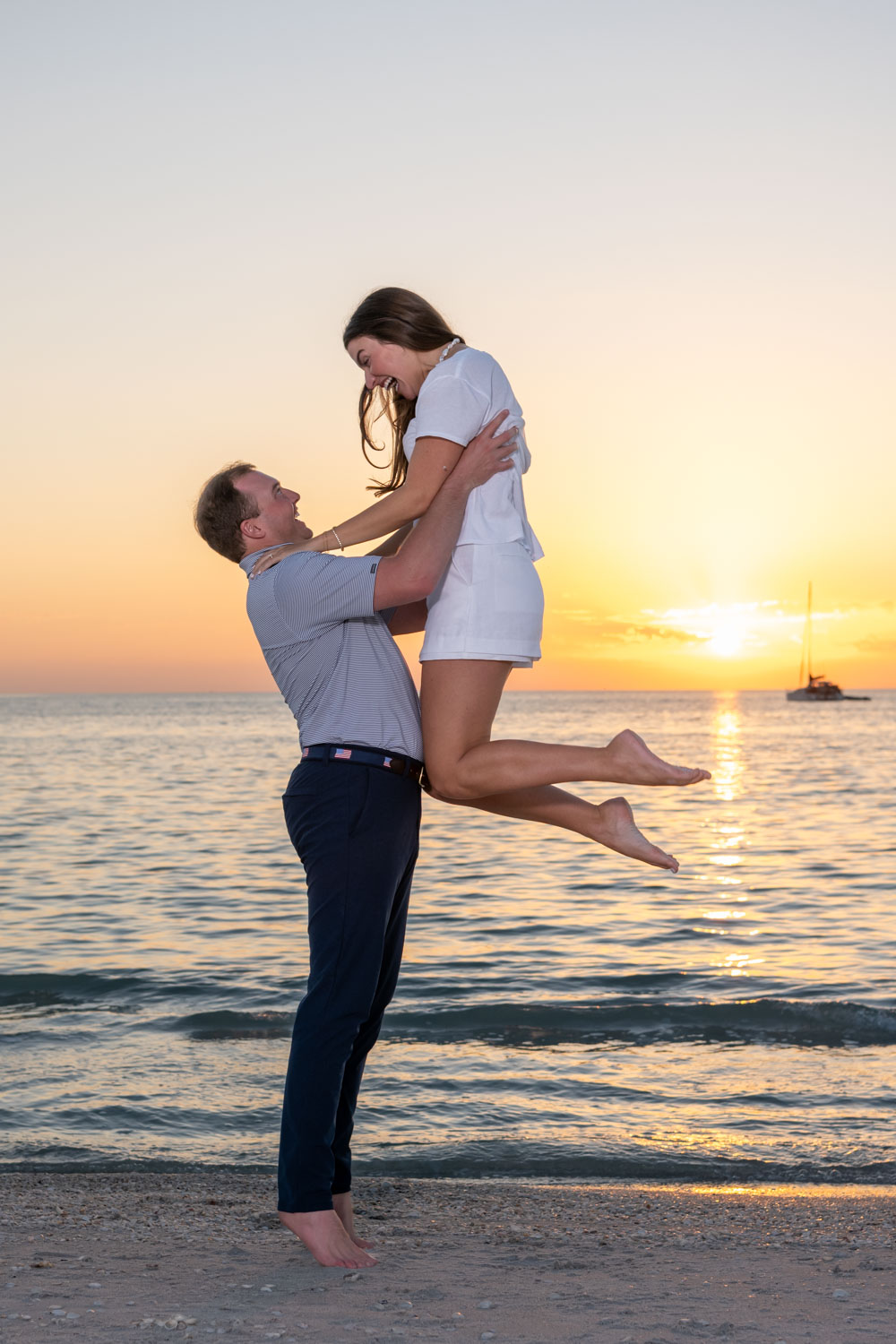 guy lifts his fiance in the air on the beach at sunset with a sailboat in the distance