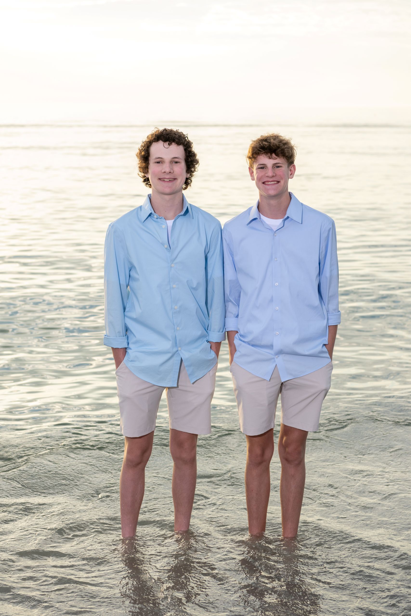 two brothers in blue shirts and tan shorts stand together in the Gulf