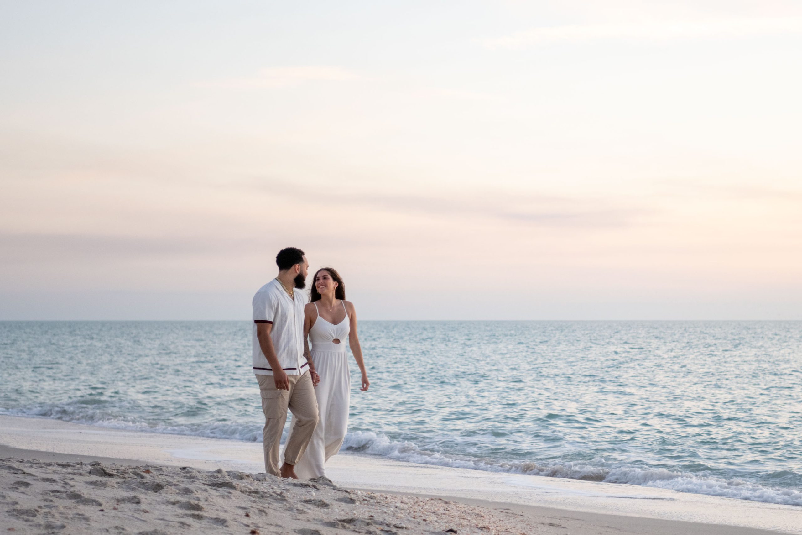couple walk along beach as they sky turns a soft pink