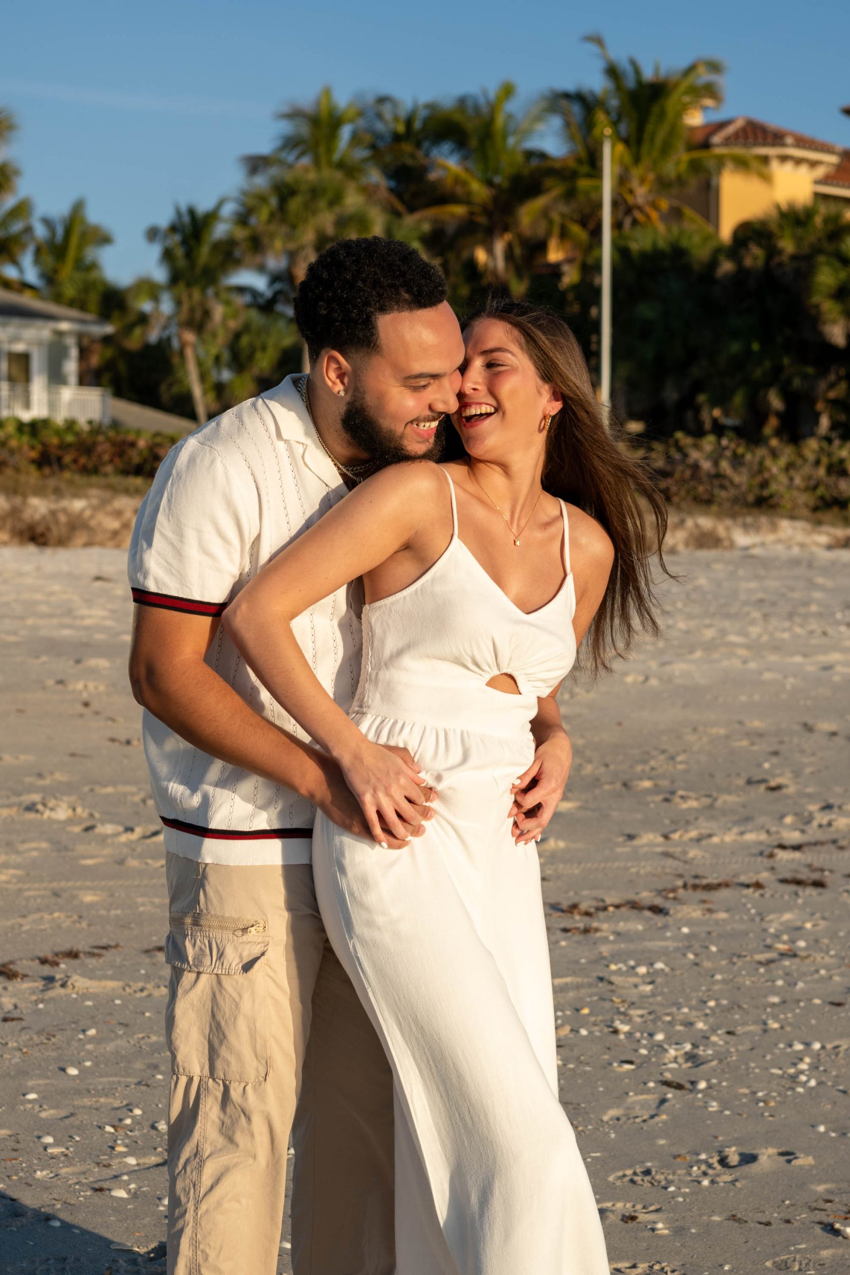engaged couple smile and laugh on the beach while he holds her from behind