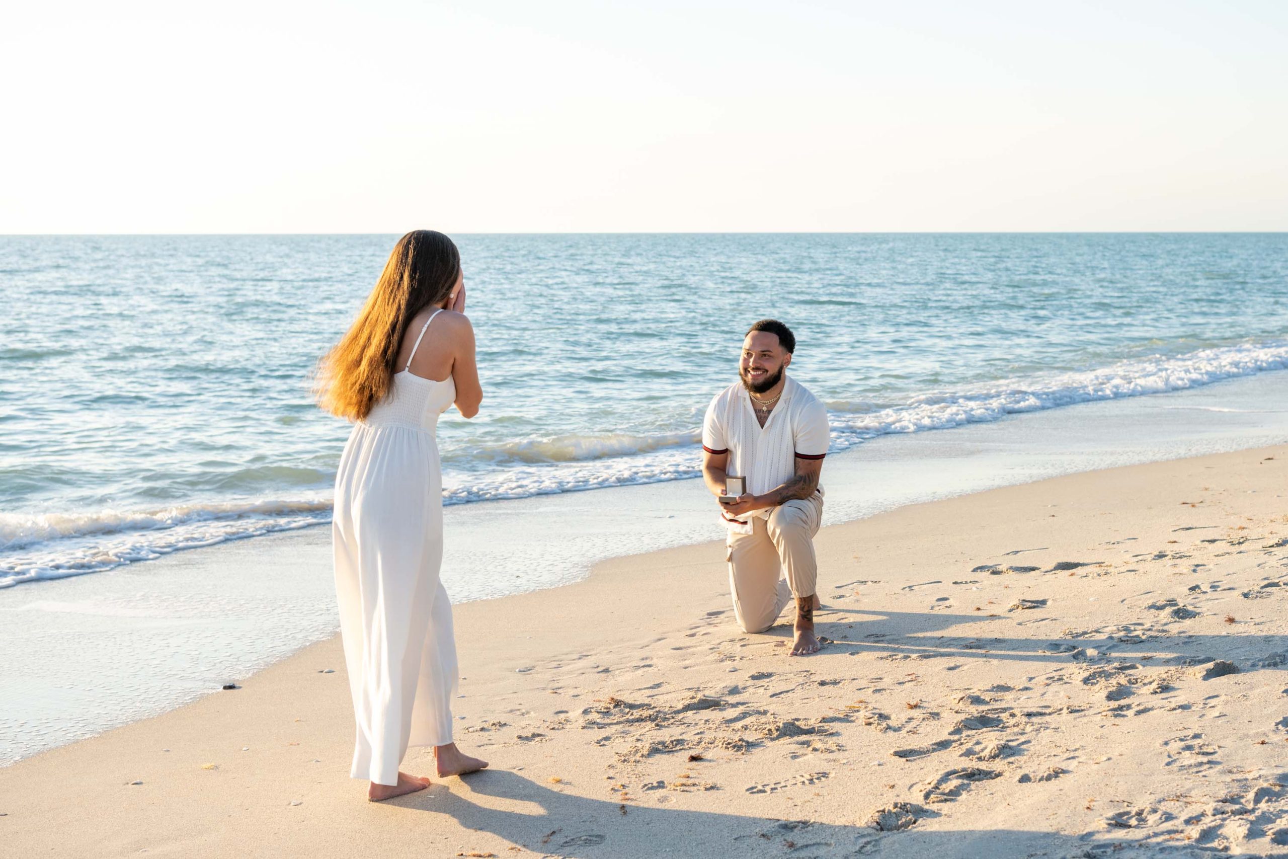 girl turns to see her boyfriend down on his knee to propose on the beach