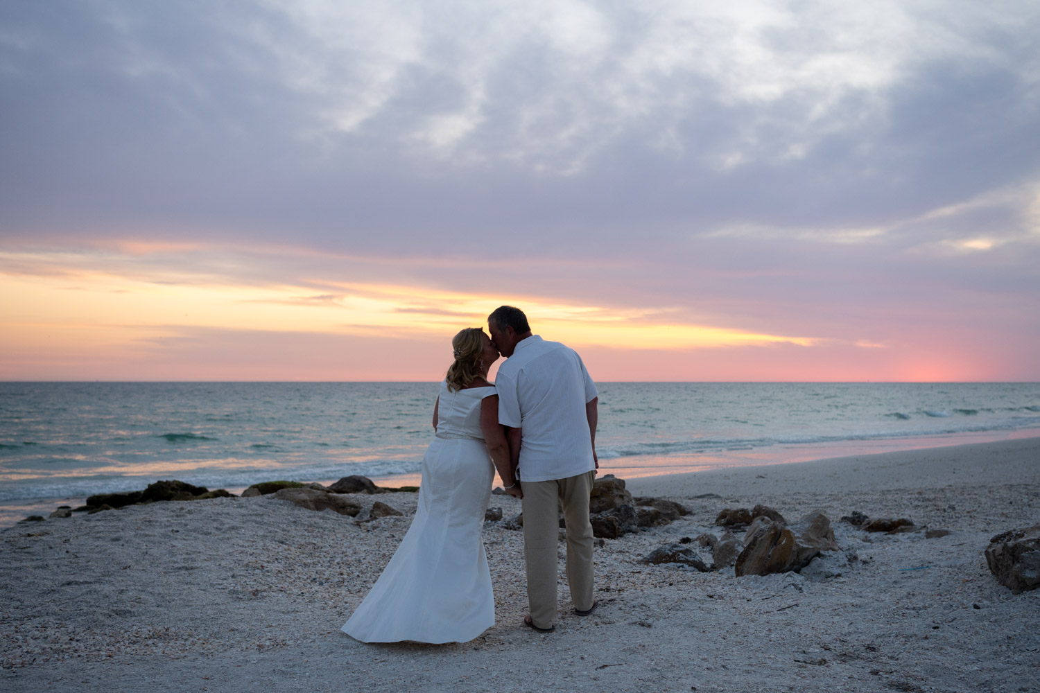 husband and wife share a kiss on the beach as the sun starts to set