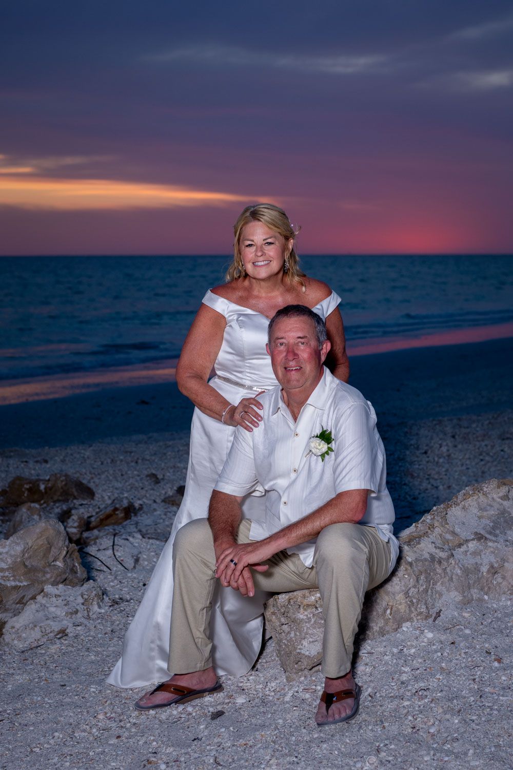 bride leans over as husband sits on rocks after wedding on Lido Beach as the sun sets