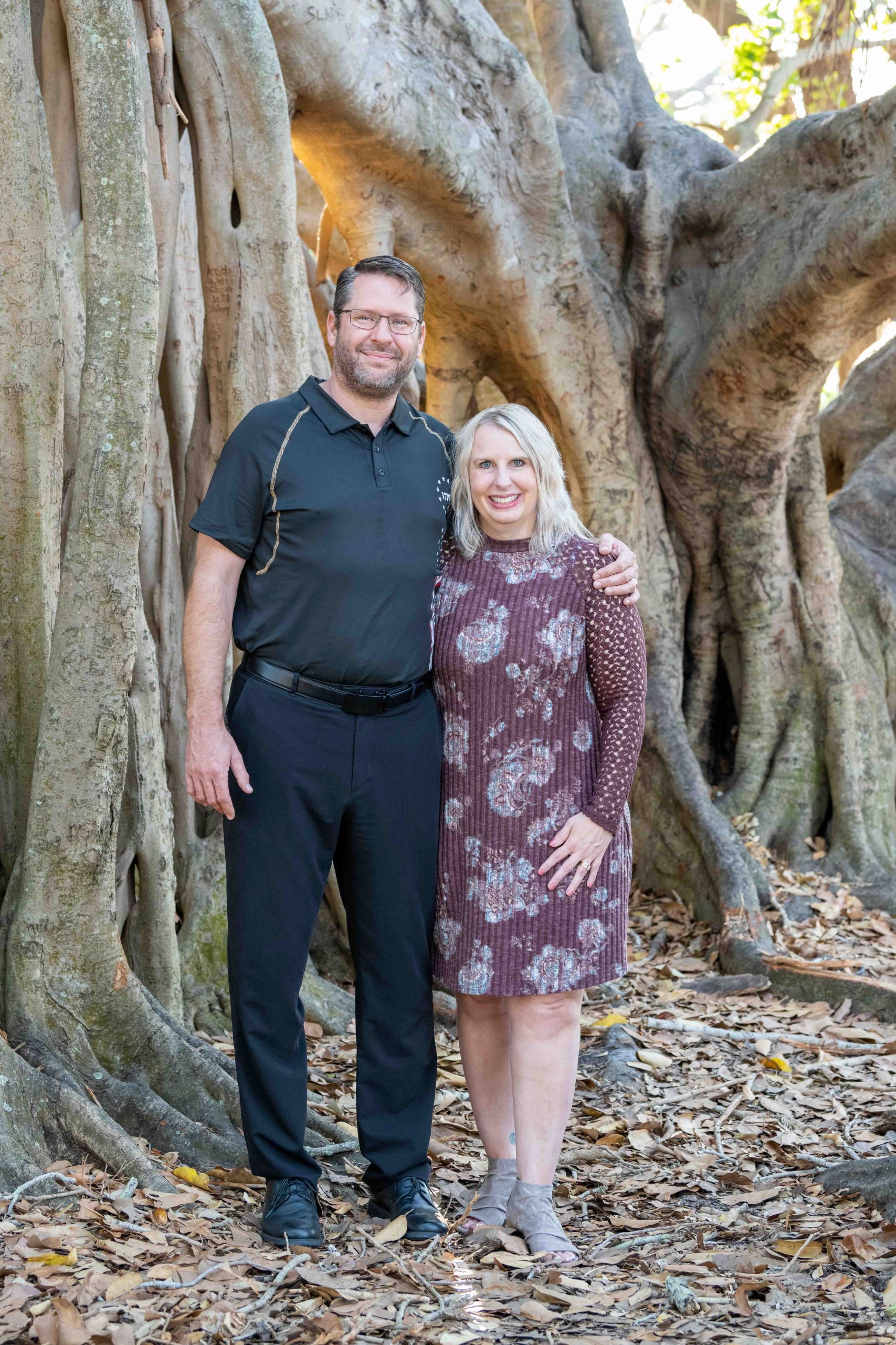 husband and wife hold each other as they stand in front of a banyan tree