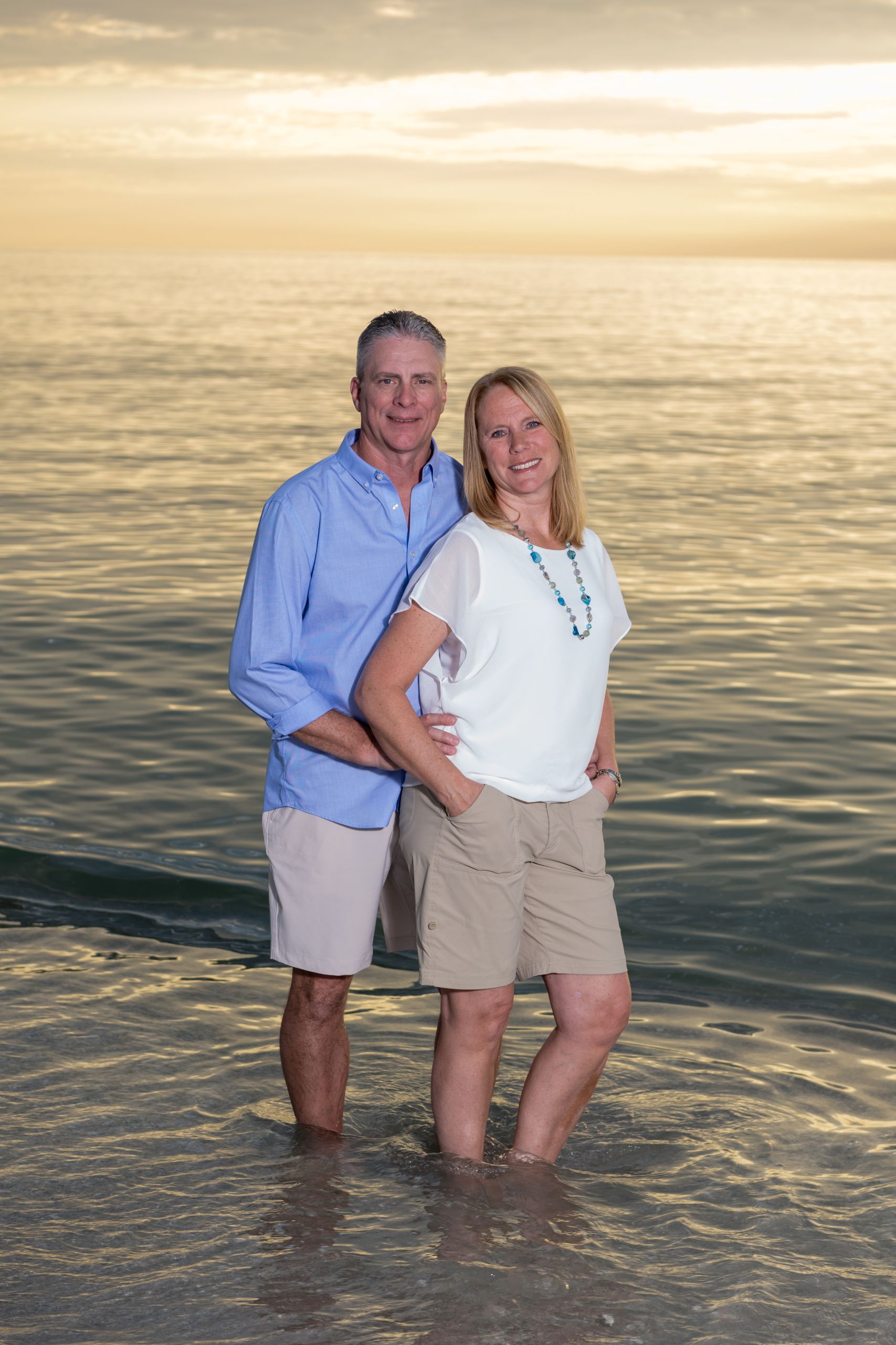 husband and wife stand in the water as the sunset turns the sky orange behind them