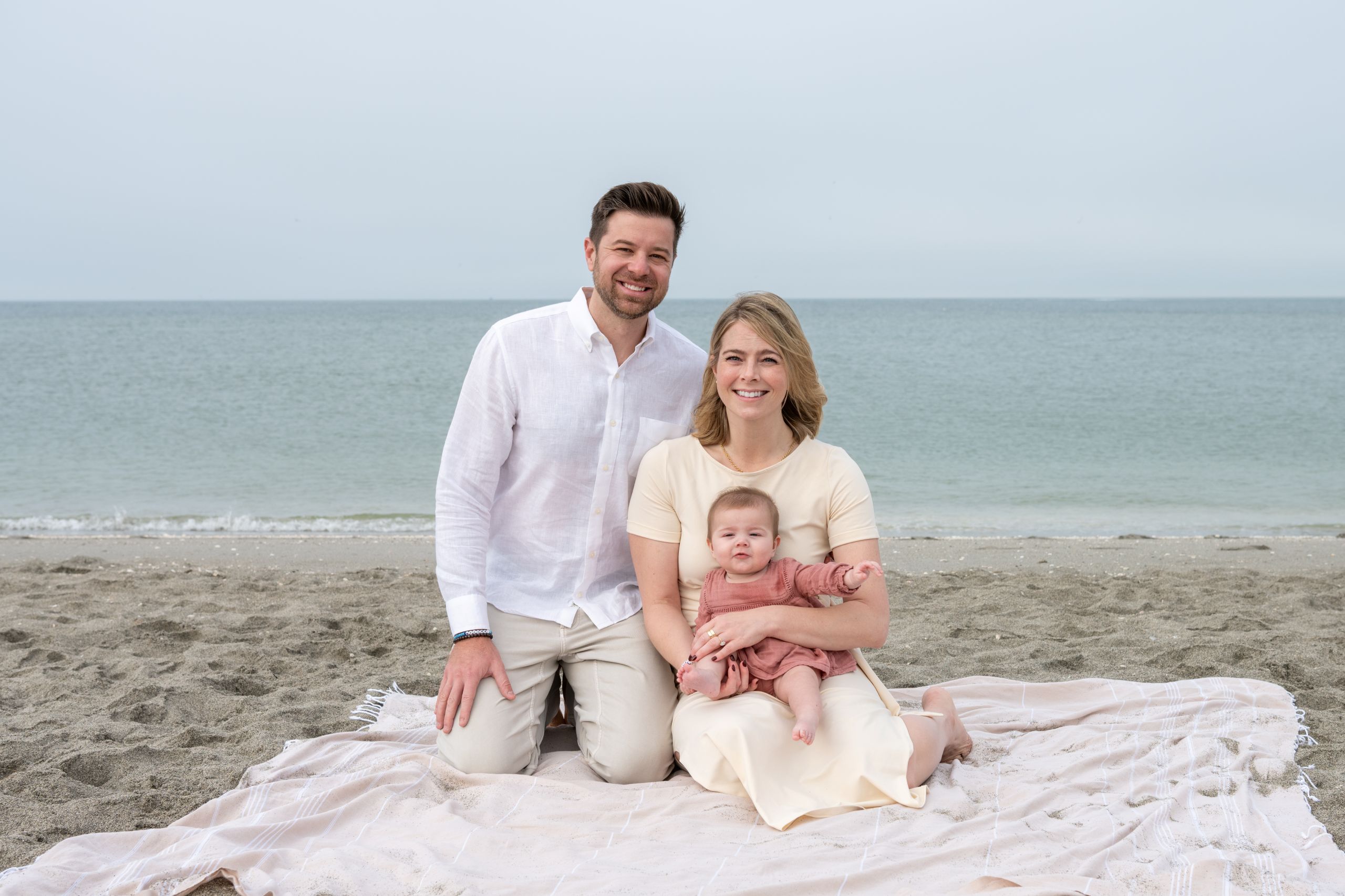 young parents with their baby girl sit on a beach blanket during morning photoshoot