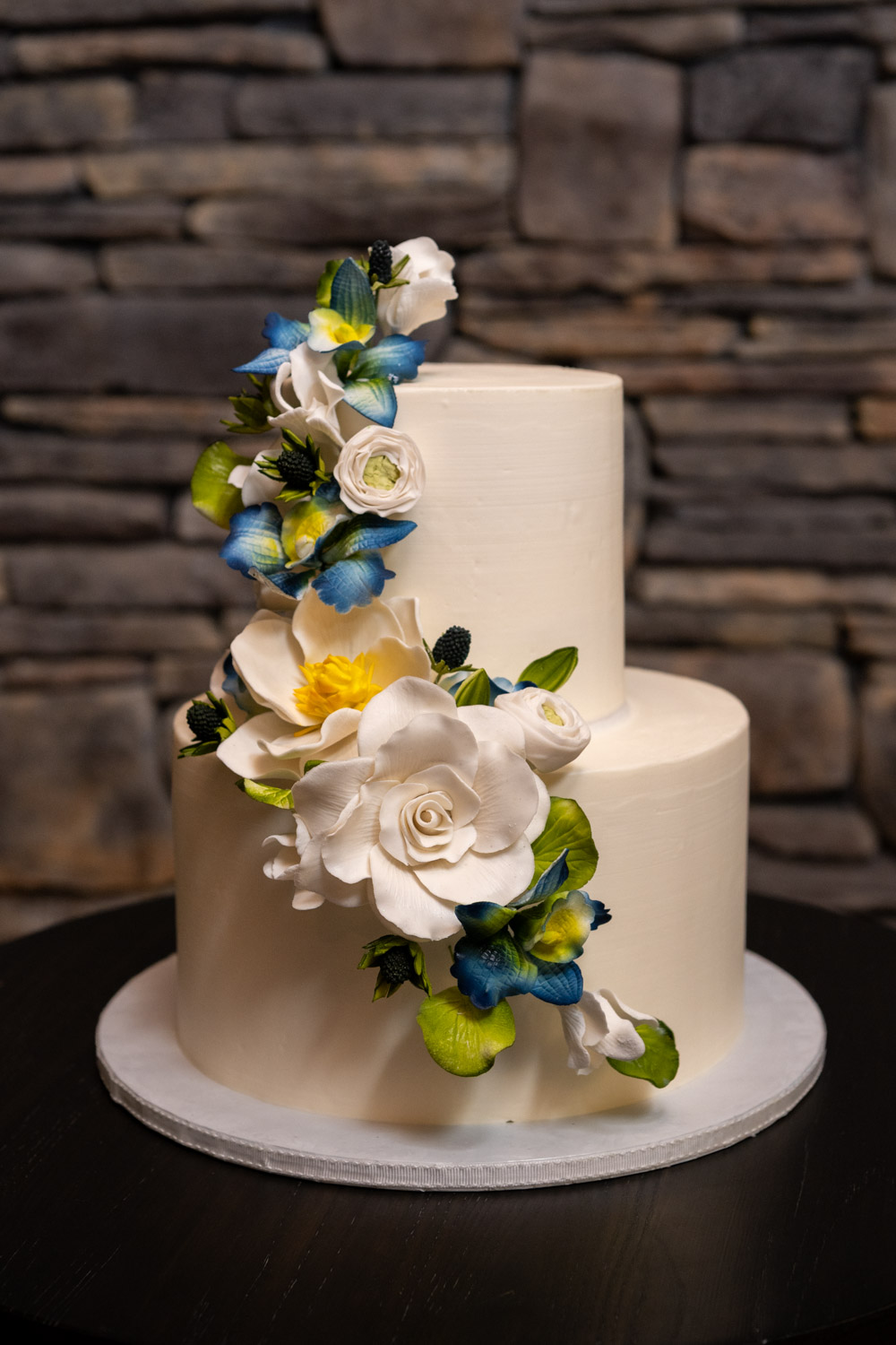 two tiered white wedding cake with white, blue and yellow flowers going up it, made by Cakes By Ron
