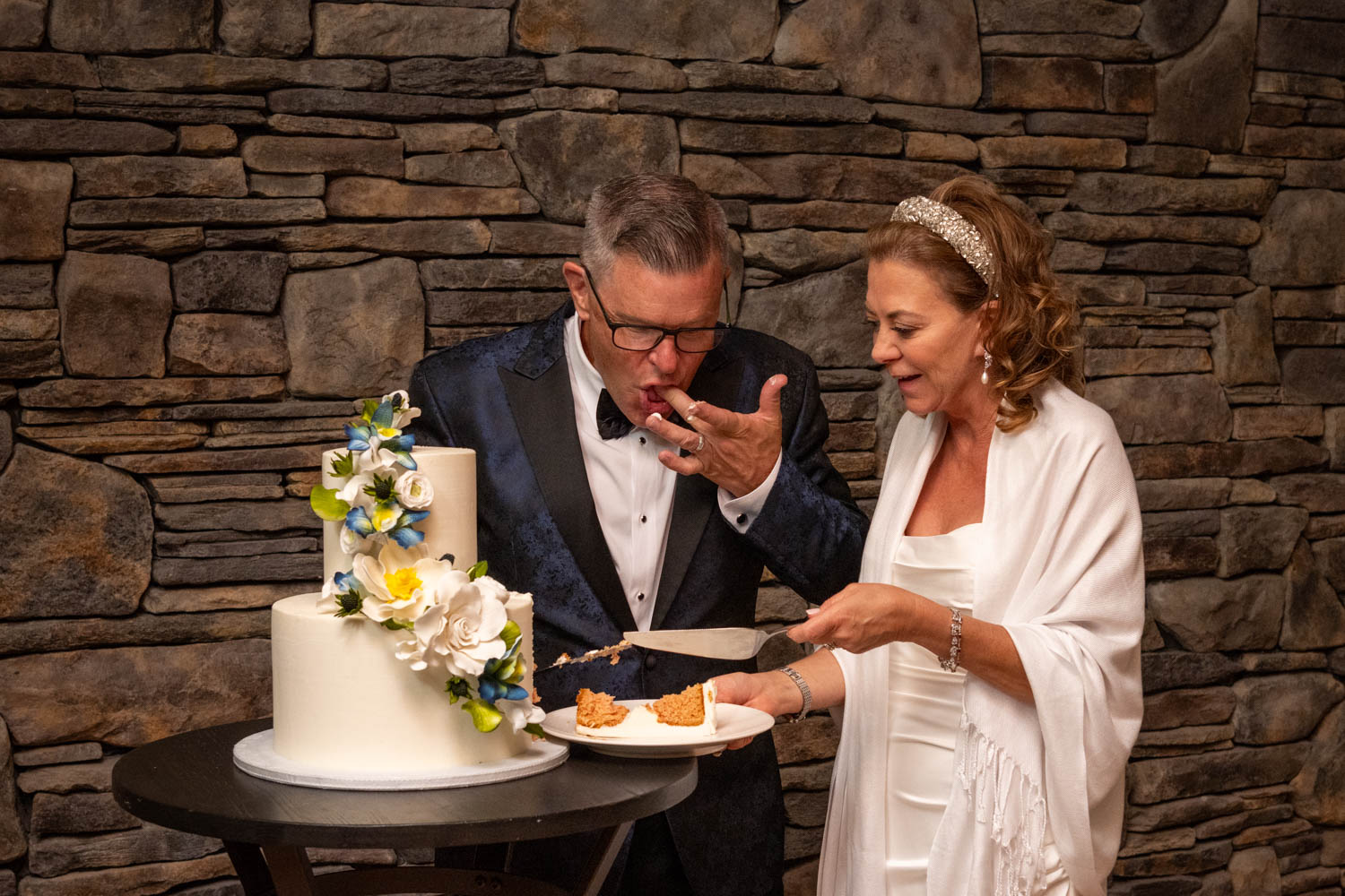 groom licks his fingers after trying the wedding cake as the bride cuts another piece