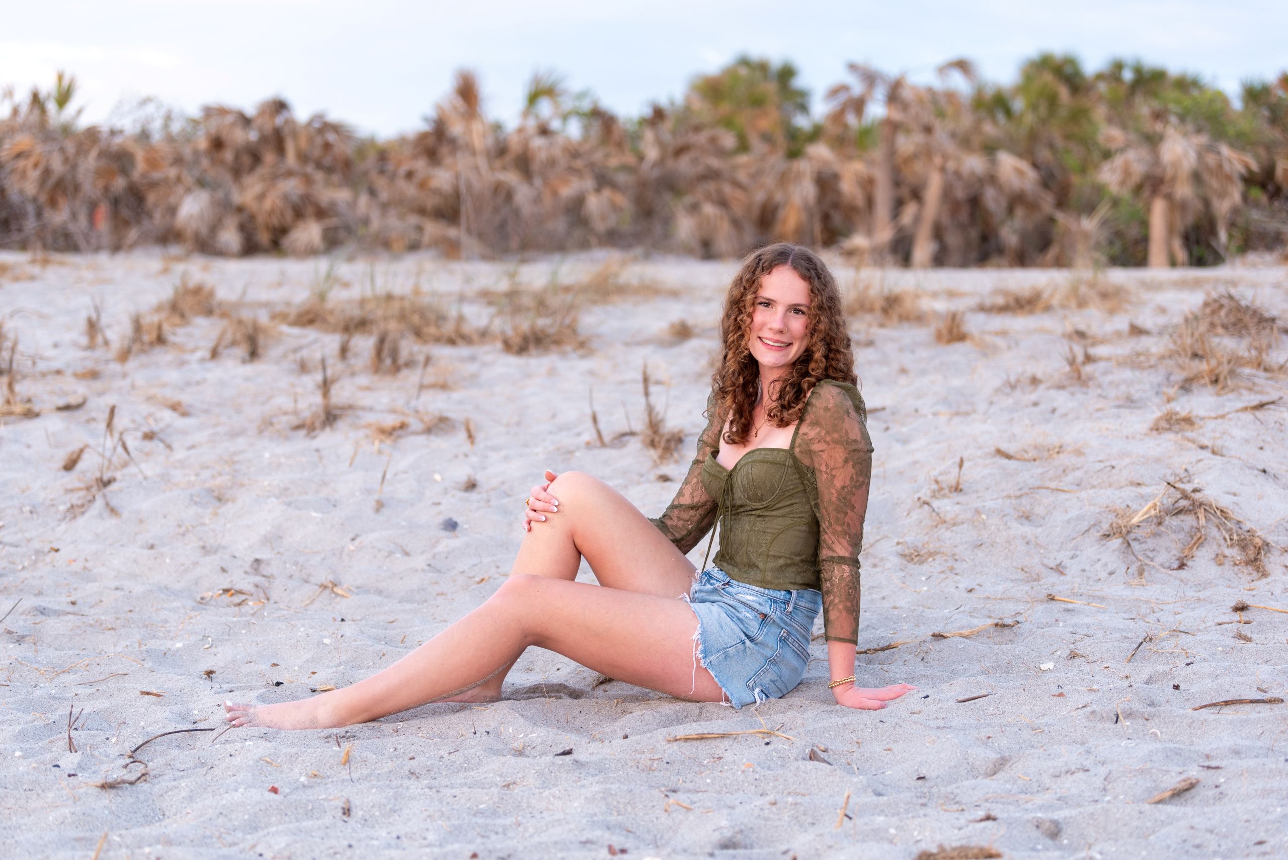 Senior girl sitting on beach in green top and blue jean shorts with sun glow on her