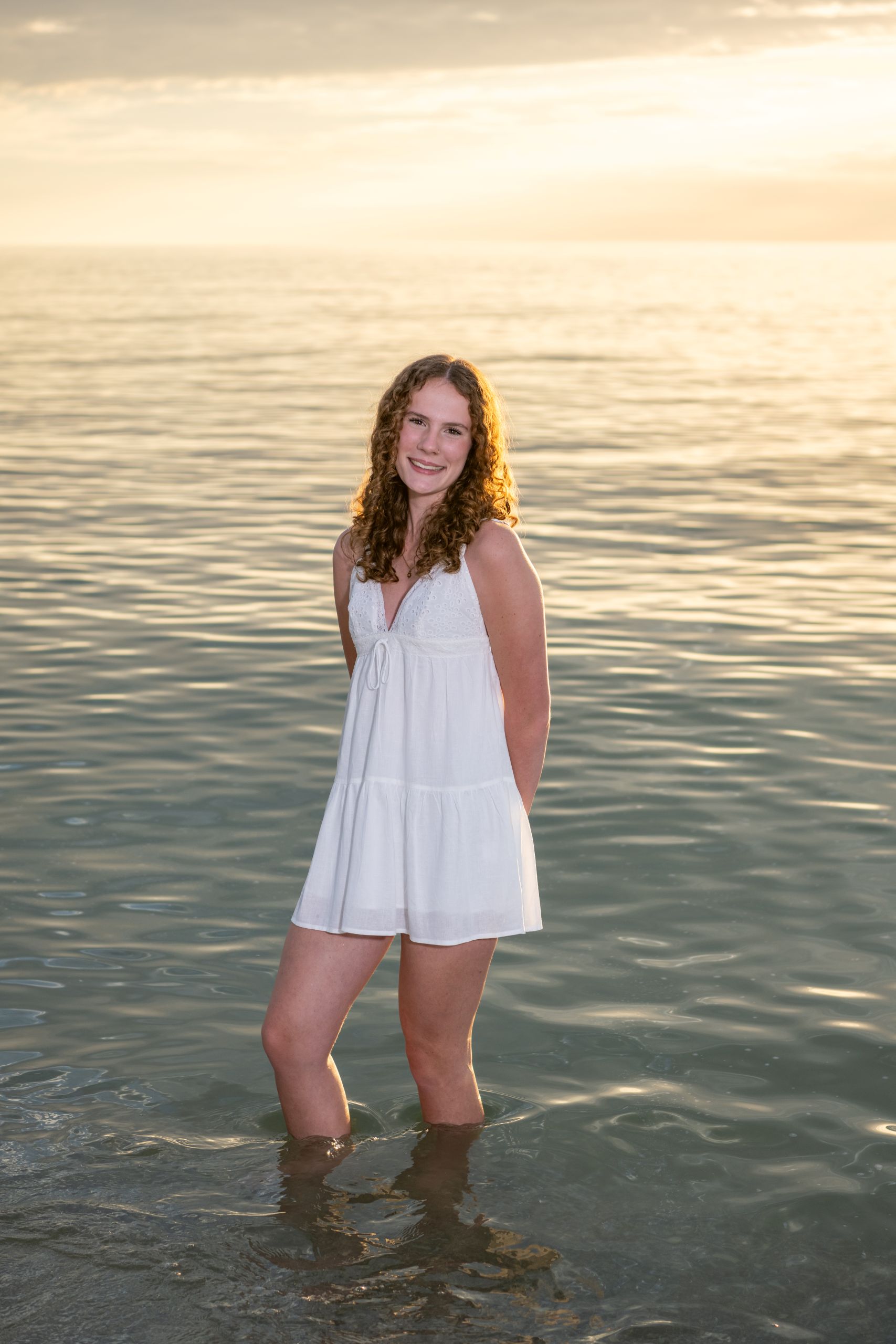 senior session with girl in white dress in the Gulf with the golden glow of sunset behind her