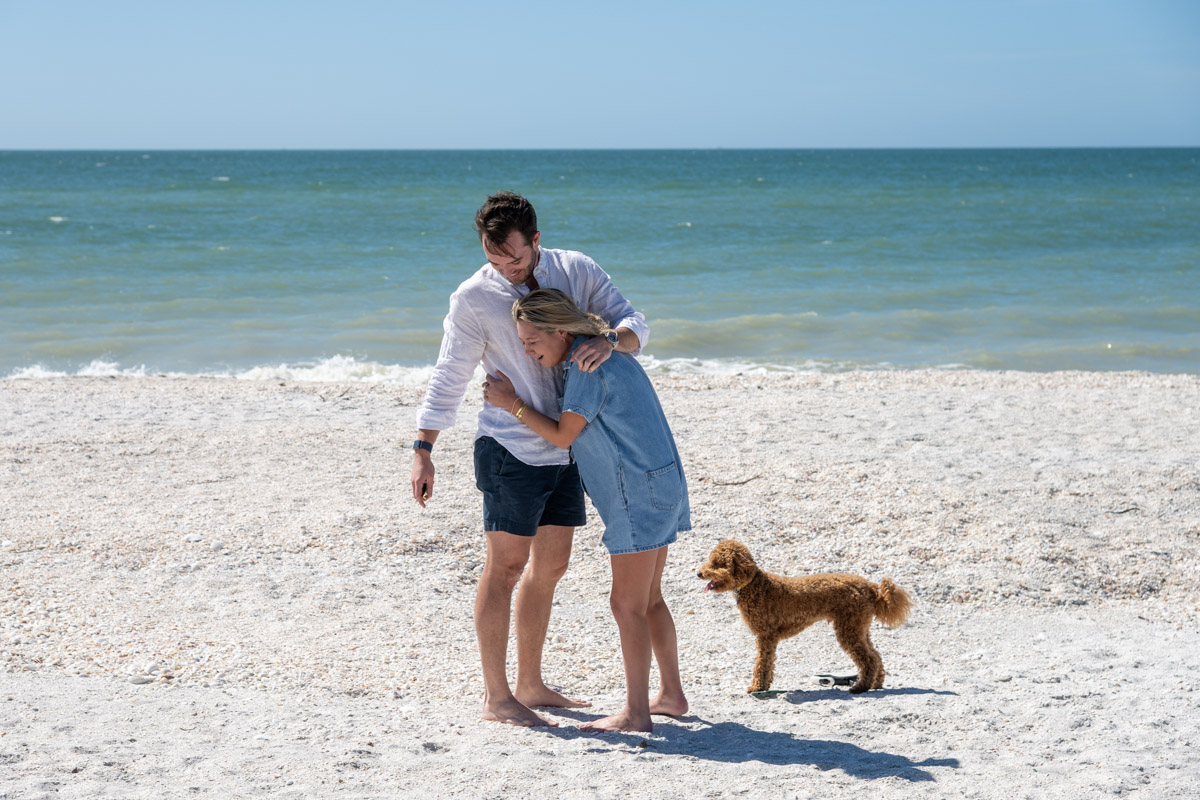 girl hugs her fiance as they stand on the beach with their dog
