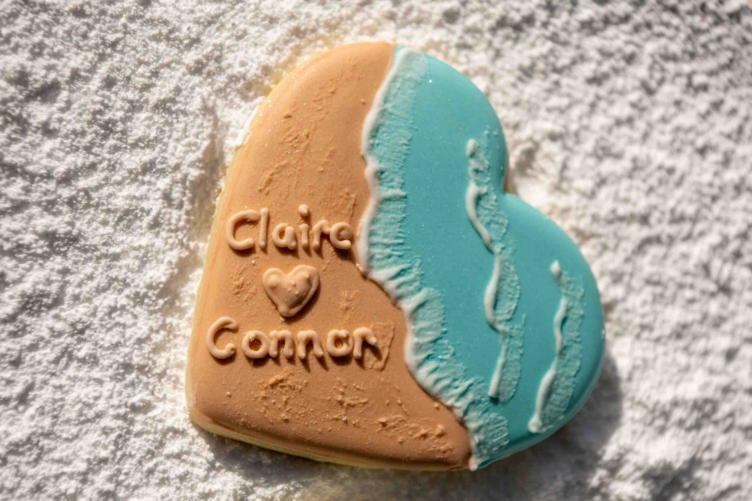 a cookie in the shape of a heart decorated with the names Claire and Connor over a beach scene