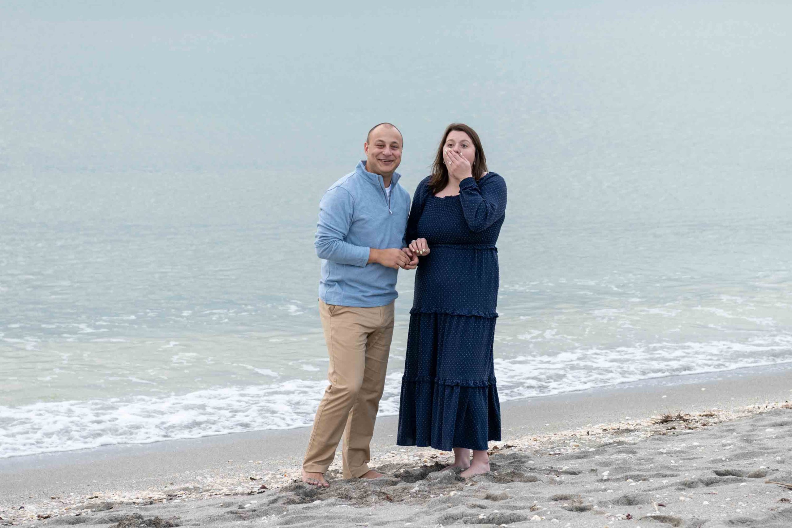 woman covers her mouth in surprise as she stands with her fiance on the beach