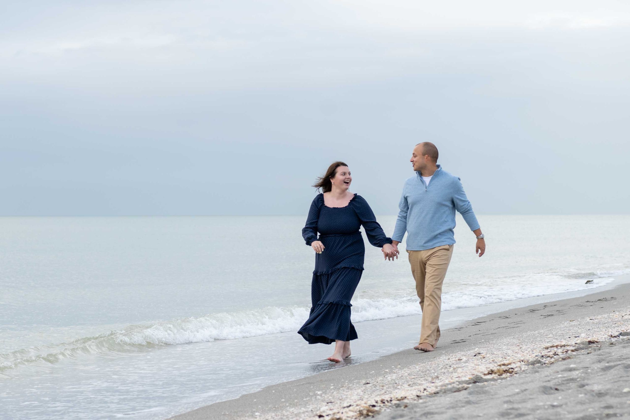 newly engaged couple walk along beach as they smile at each other