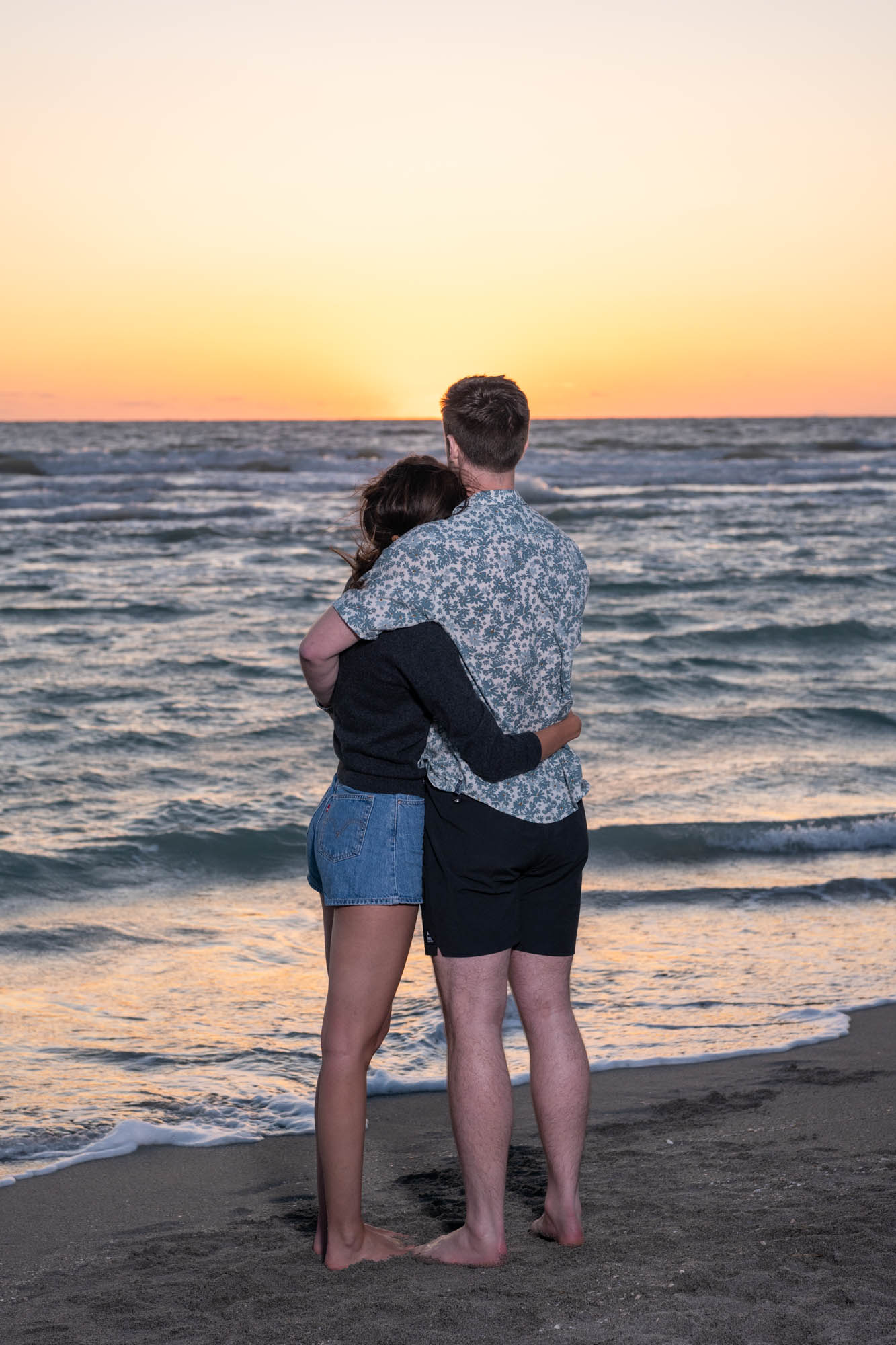 newly engaged couple hold each other as they watch the sunset on the beach