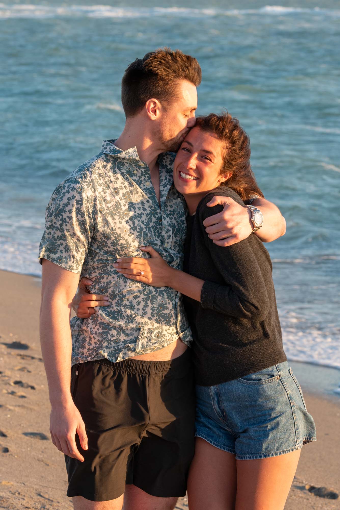 man kisses his fiance as he hugs her after proposing on the beach