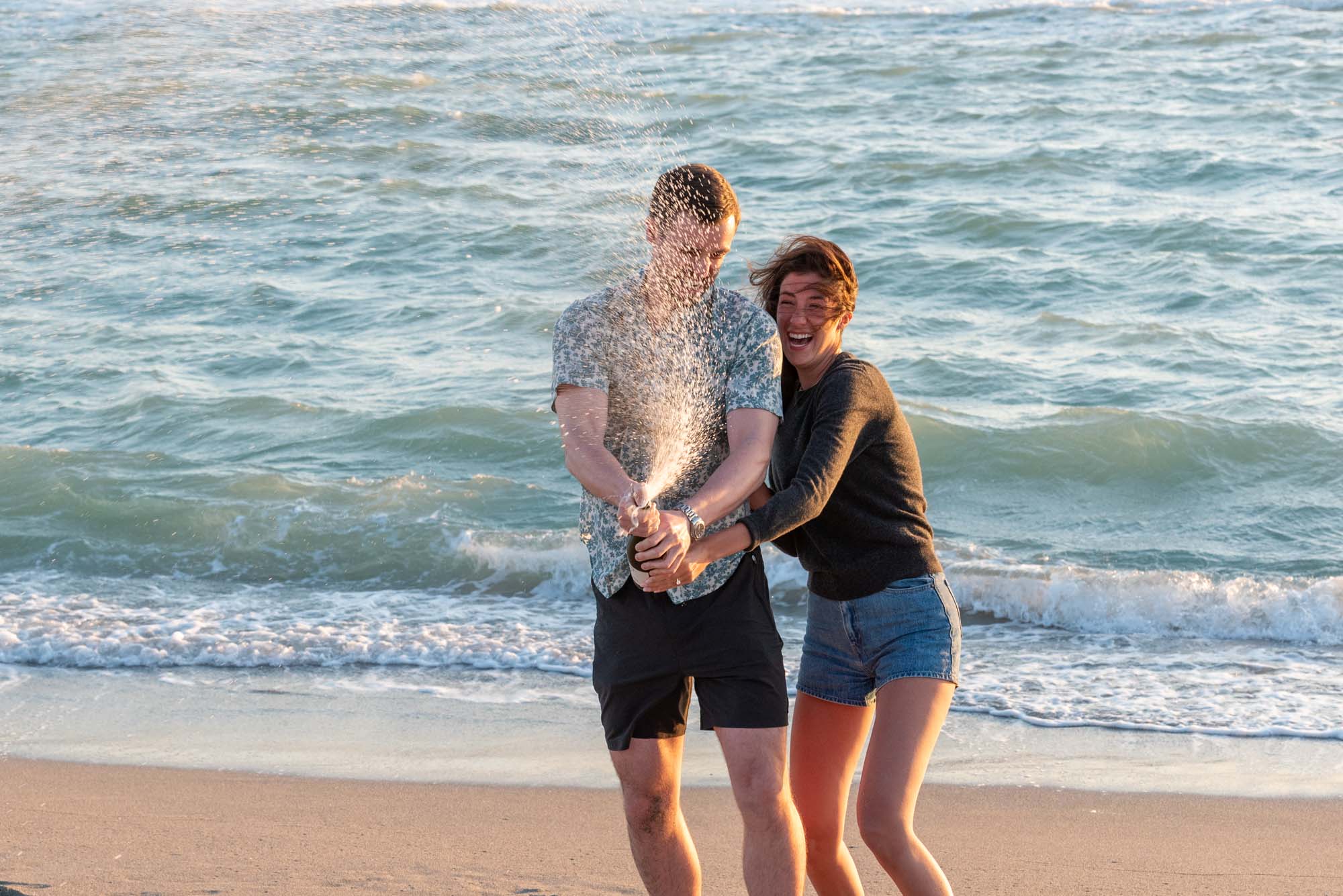 couple laugh as they spray champagne on the beach