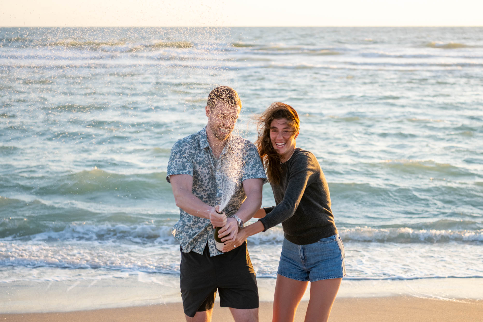 newly engaged couple pop a bottle of champagne on the beach in celebration and they laugh as it sprays everywhere