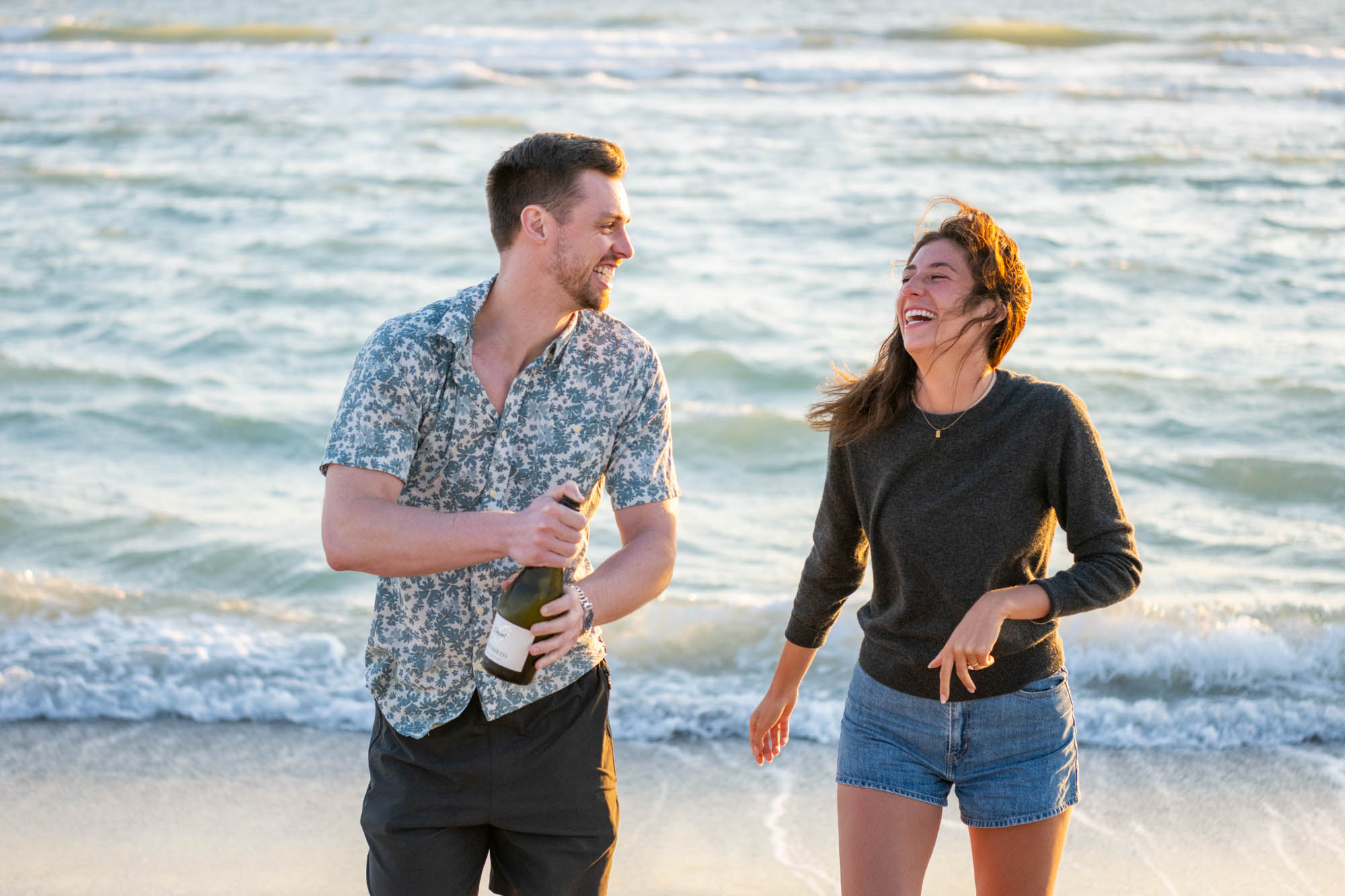 couple laugh after popping champagne on the beach