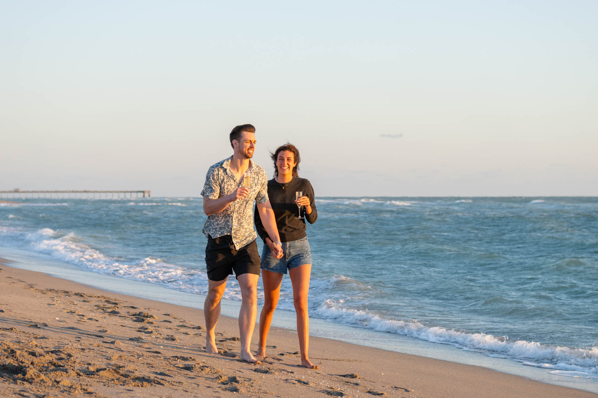 couple walk along Venice Beach with champagne glasses