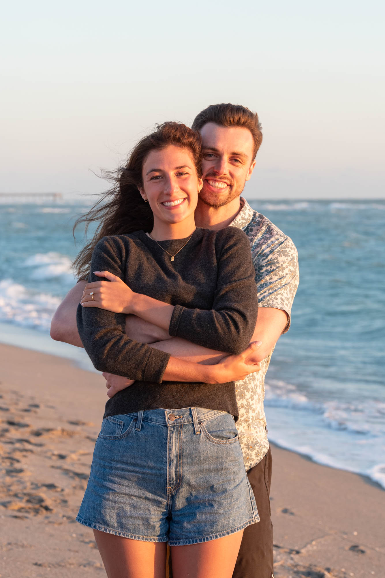 man hugs his fiance from behind while standing on beach