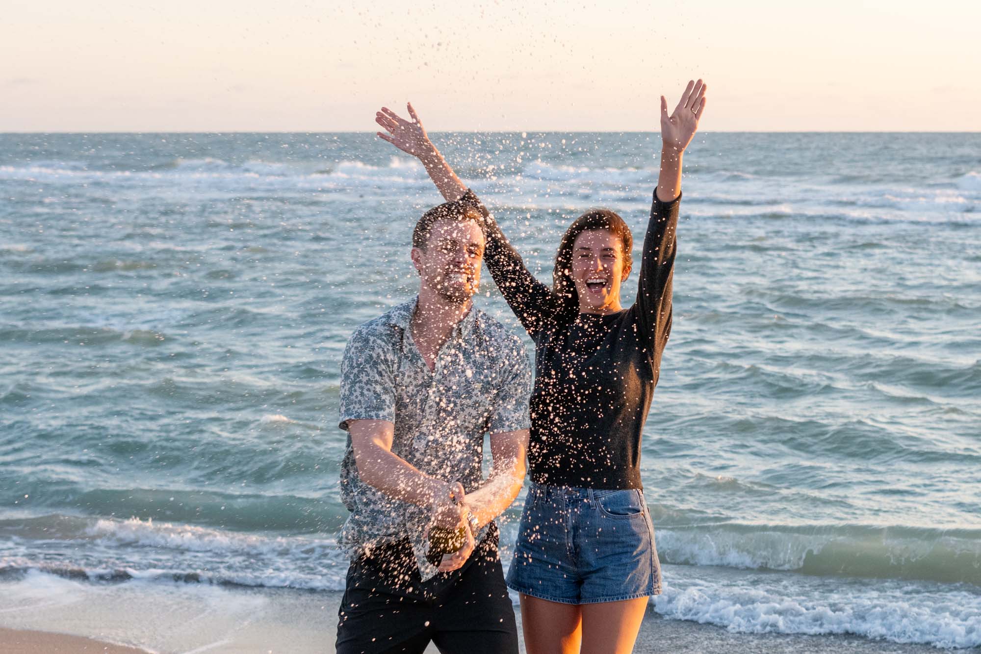 girl cheers as her fiance sprays champagne as the celebrate on the beach