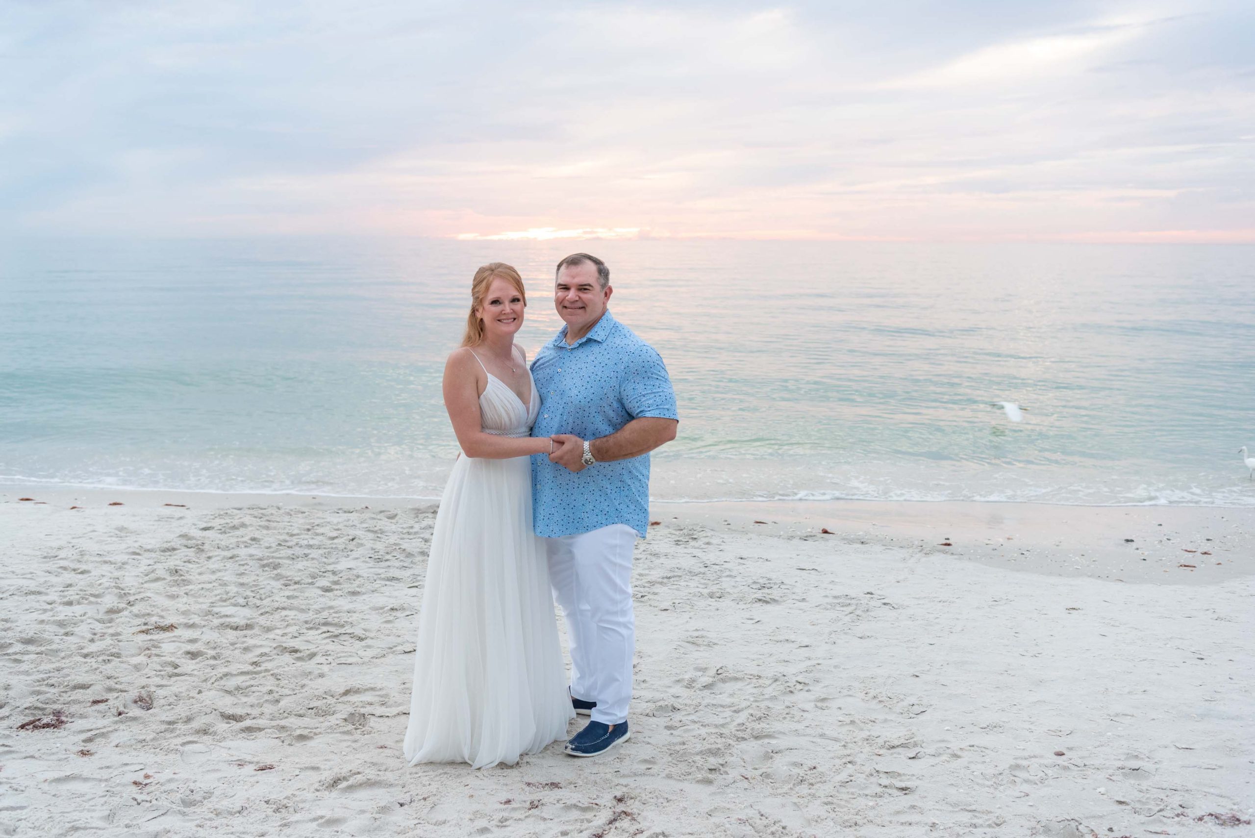 bride and groom hold each other close on white sand beach as the sun gently sets behind them