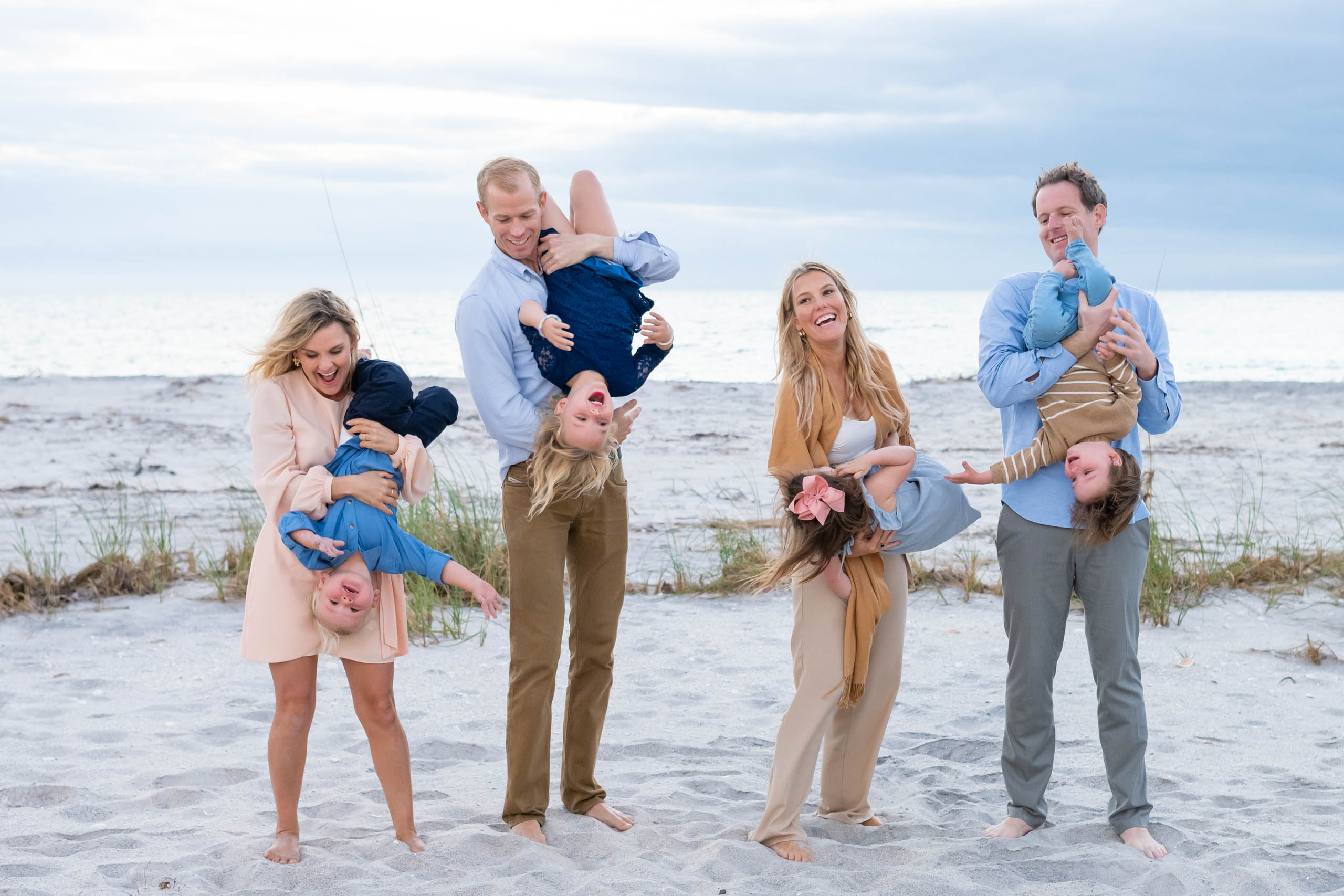 four parents twirl their children making them laugh as they stand on the beach with the sand and Gulf behind them