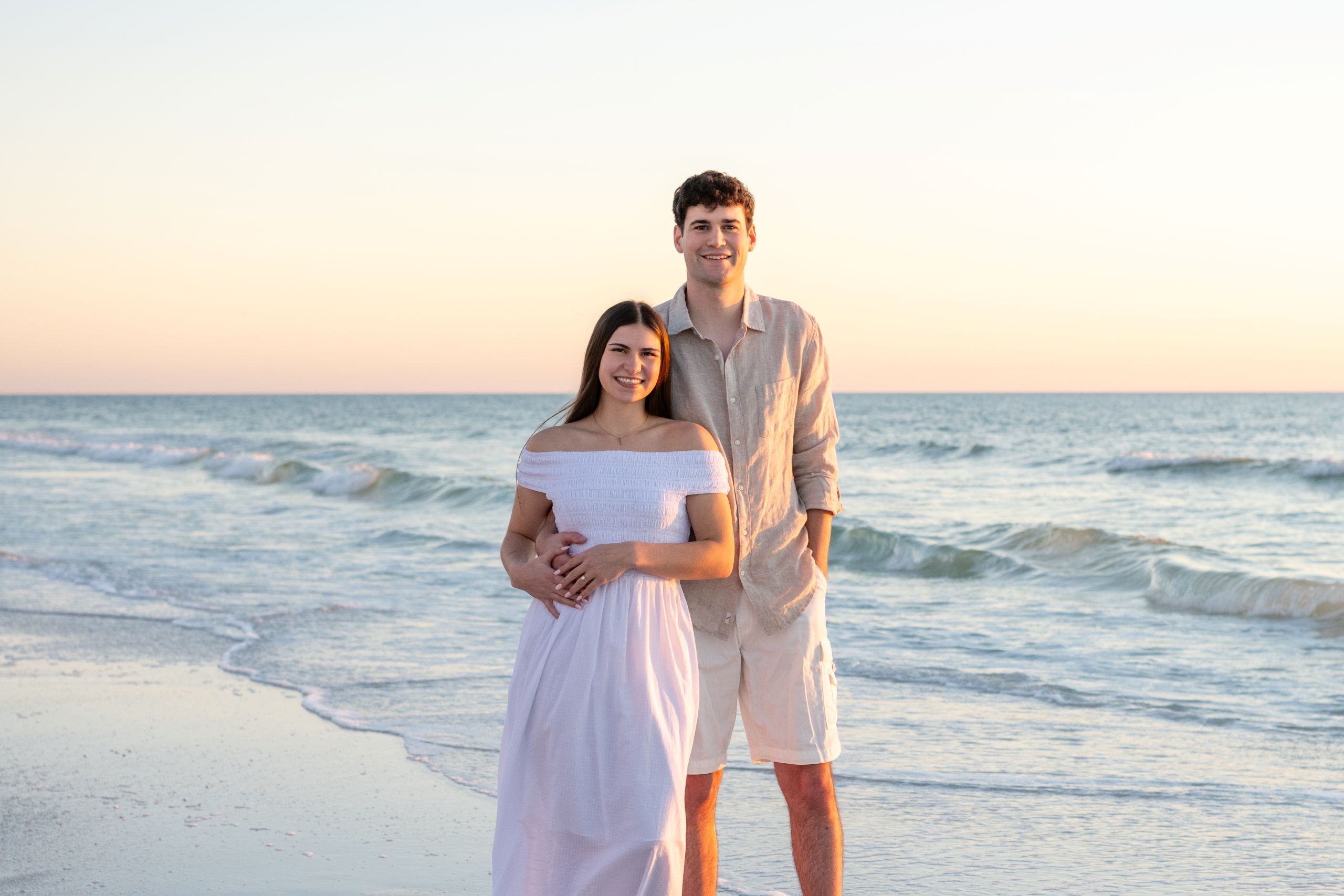 engaged couple hold each other as they stand on the beach with the golden hour glow of sunset behind them