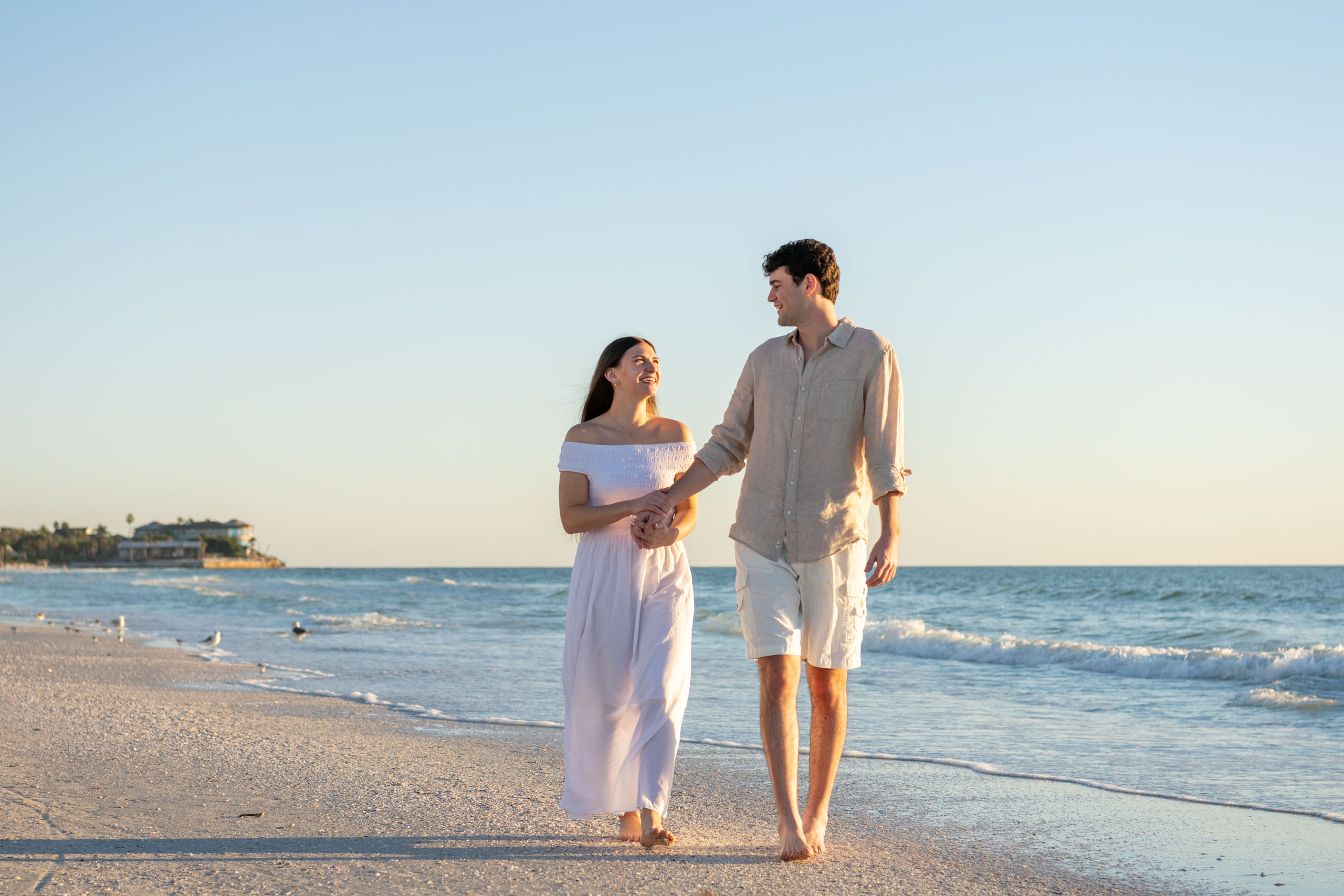 engaged couple walk along beach holding hands smiling at each other