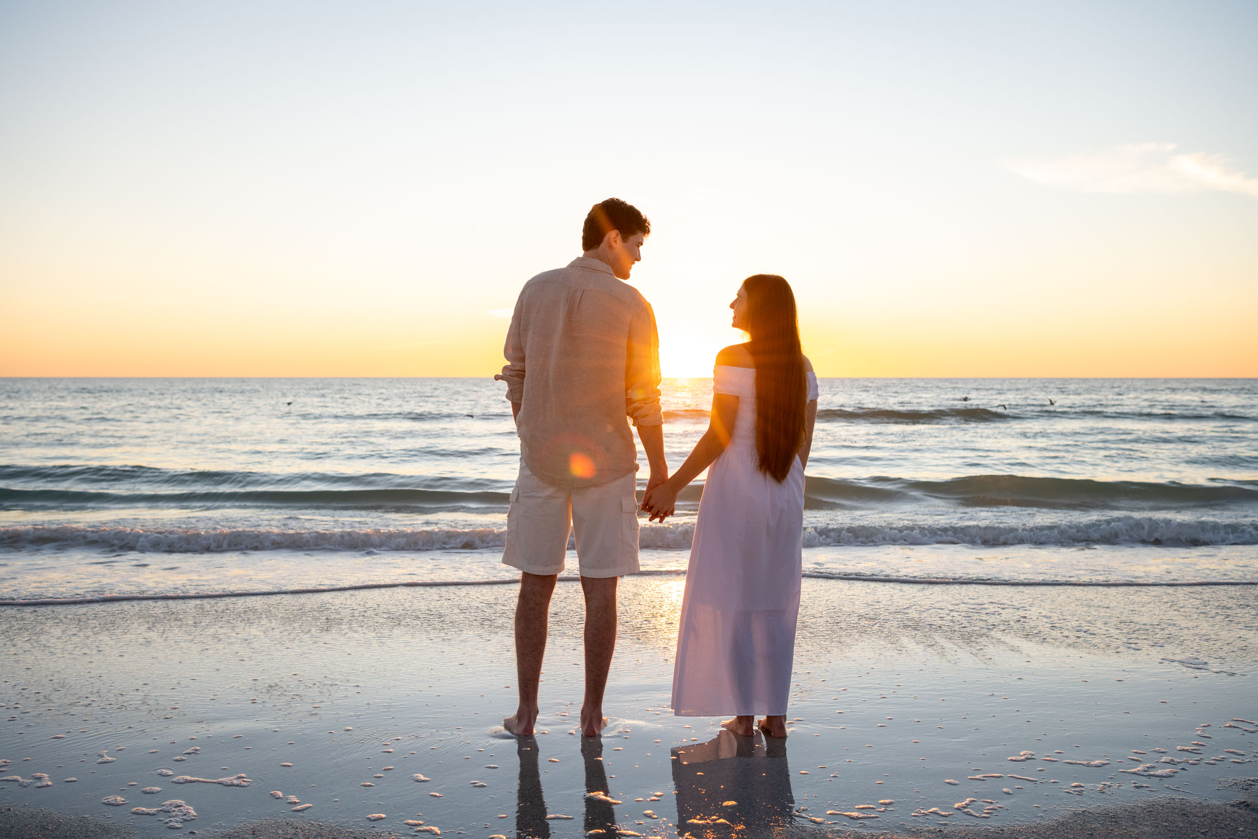 couple holding hands, looking at each other as they stand on the beach