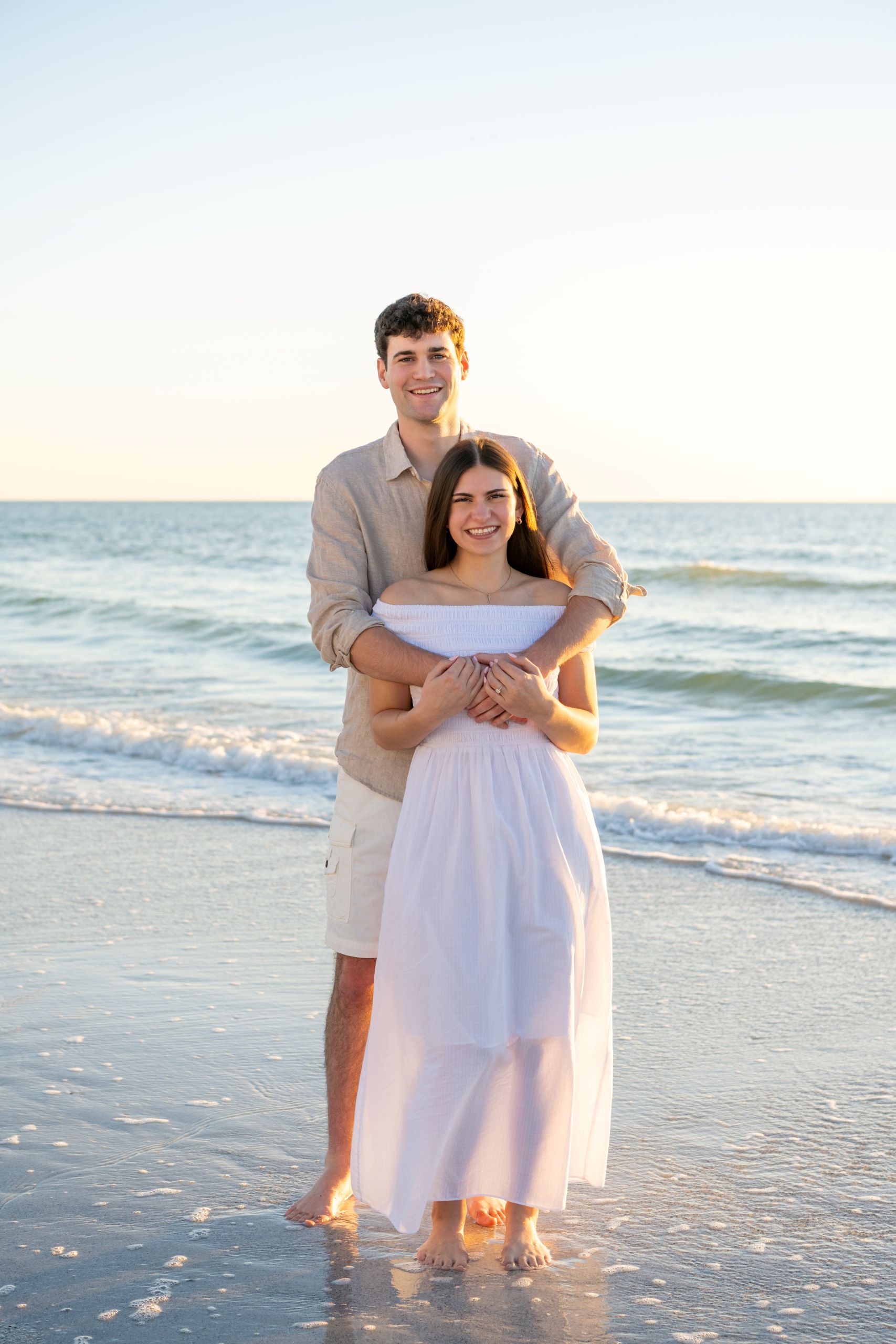 man wraps his arms around his fiance as they stand on the beach